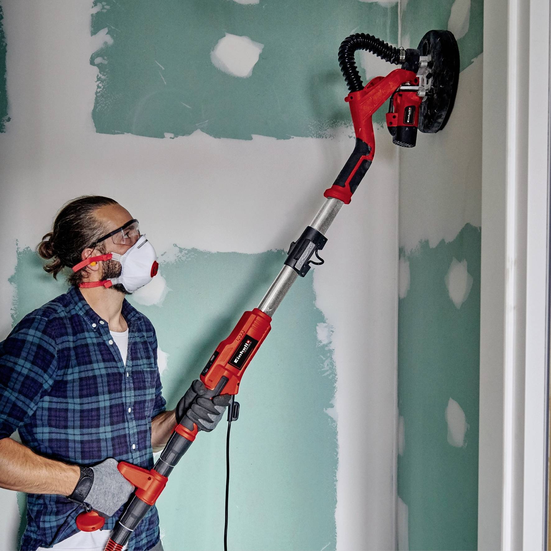 Person schleift eine Wand mit einer elektrischen Schleifmaschine. Sie trägt Schutzbrille und Atemmaske. Hintergrund: unverputzte Wand.