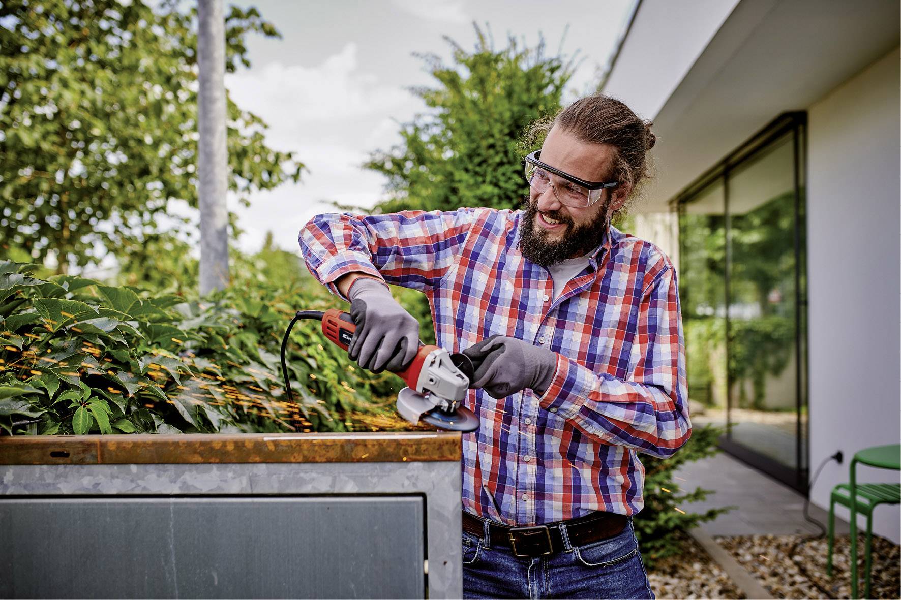 Eine Person mit Schutzbrille und Handschuhen schneidet Äste mit einer Motorsäge in einem Garten. Sie trägt ein kariertes Hemd.