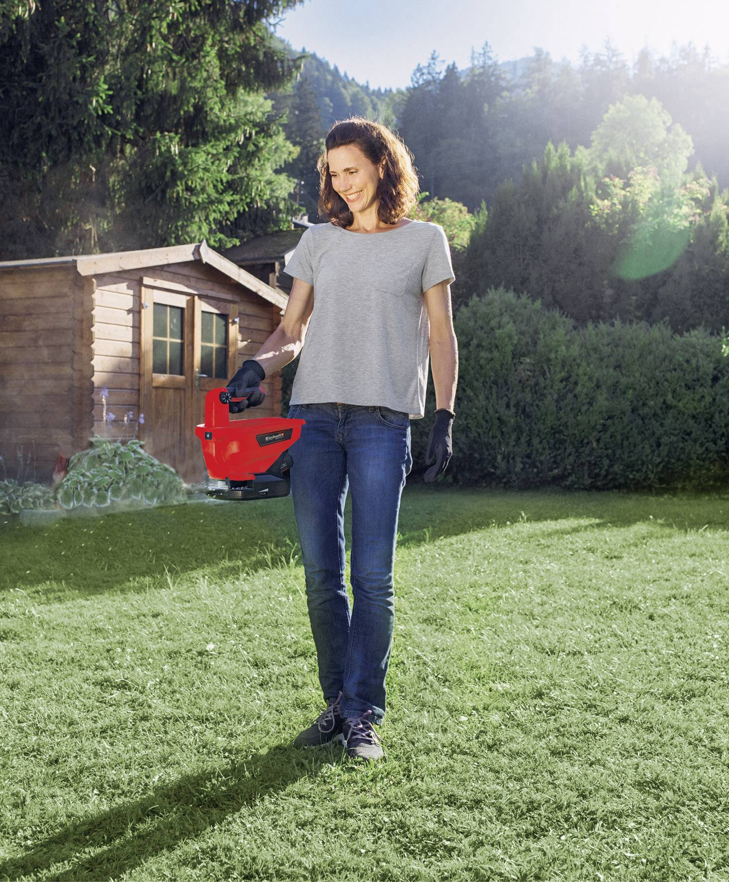 Eine Frau steht im Garten mit einem roten Rasenmäher in der Hand, umgeben von grünen Bäumen und einem Holzschuppen im Hintergrund.