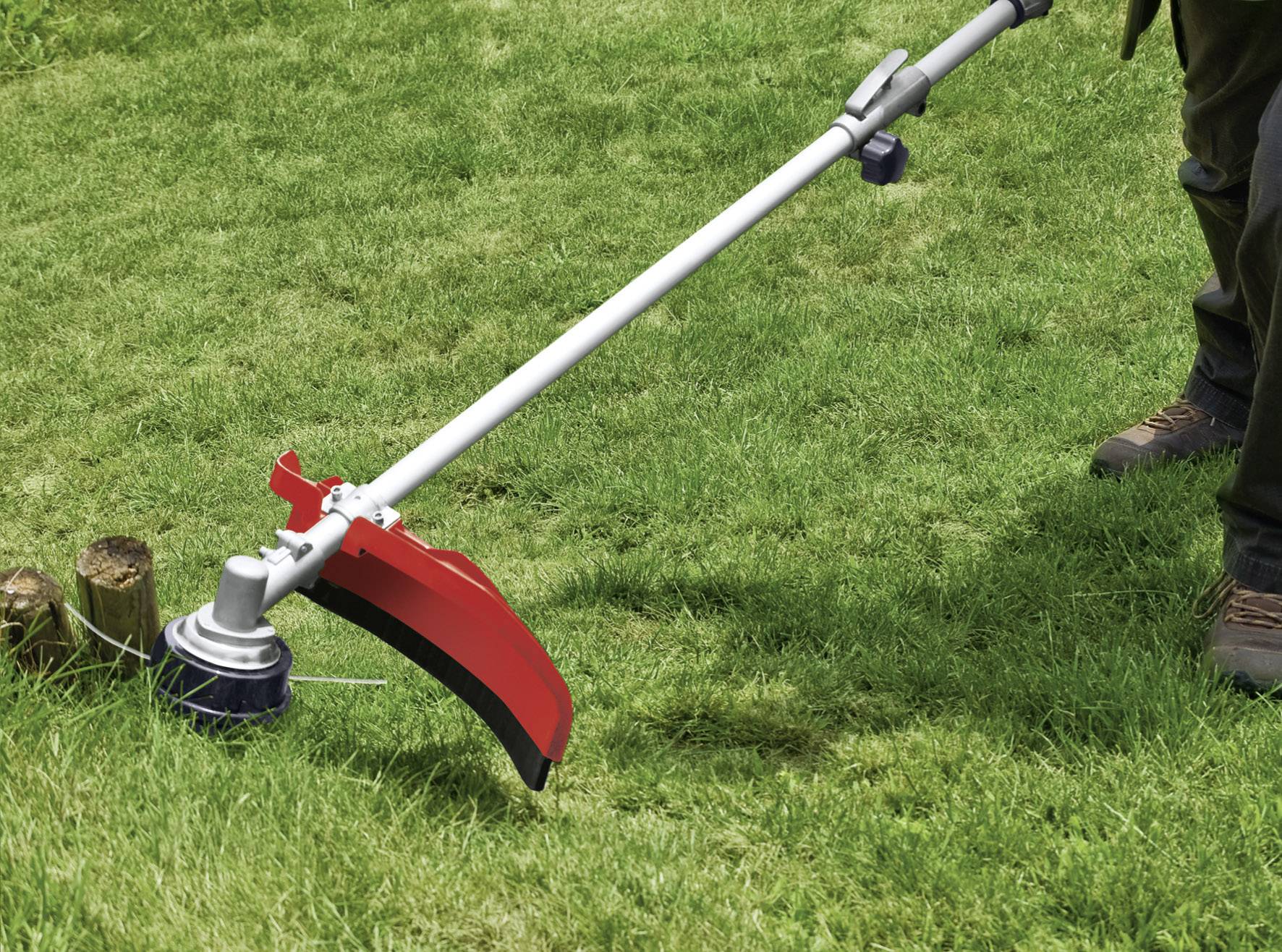 Eine Person schneidet Gras mit einem Motorsense-Trimmer auf einer grünen Rasenfläche. Ein roter Schutzschild ist am Trimmer befestigt.