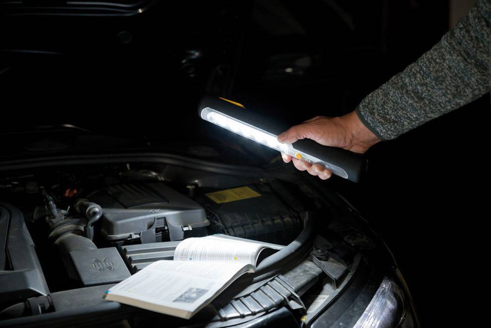 Person hält Taschenlampe über geöffnetem Motorhaube eines Autos. Ein Handbuch liegt auf dem Motor, das Licht beleuchtet den Bereich.