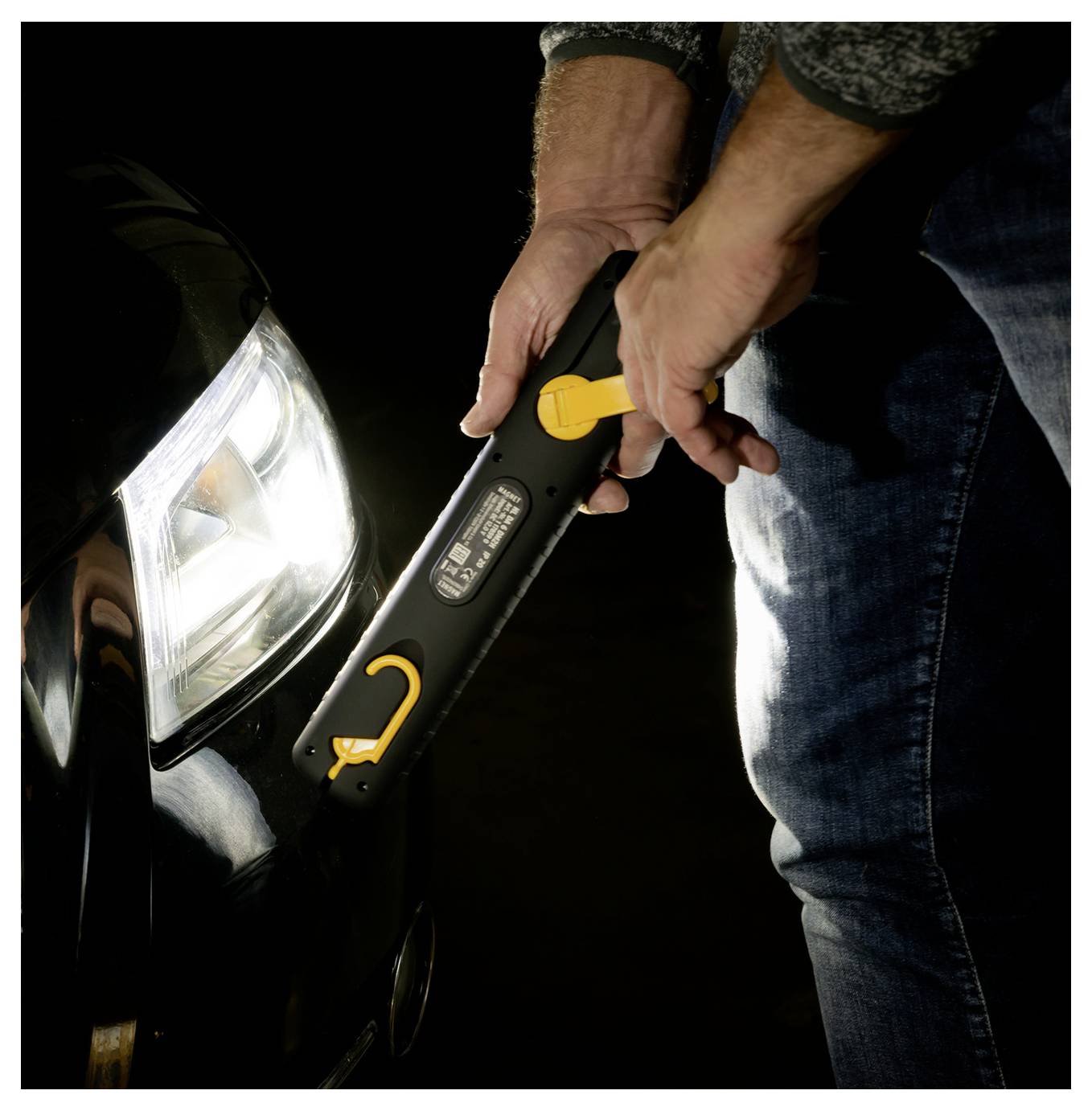 Person, der einen tragbaren Starthilfegerät an einer Autobatterie in der Nähe des vorderen Scheinwerfers in einer dunklen Umgebung benutzt.