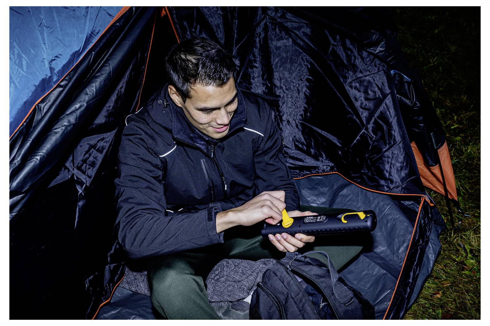 Ein Mann in einem Zelt benutzt nachts ein tragbares Gerät, wahrscheinlich um Campingausrüstung einzustellen oder aufzupumpen.