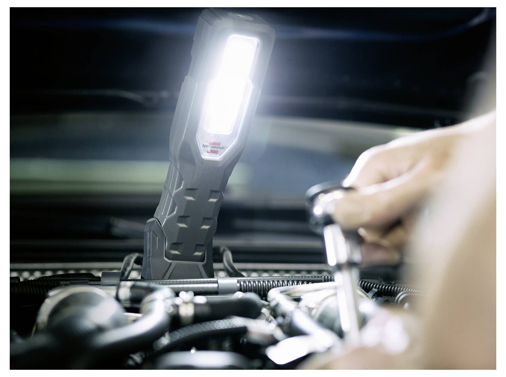 Tragbare Arbeitsleuchte, die einen Motorraum eines Autos beleuchtet, während ein Mechaniker einen Schraubenschlüssel im Vordergrund verwendet.