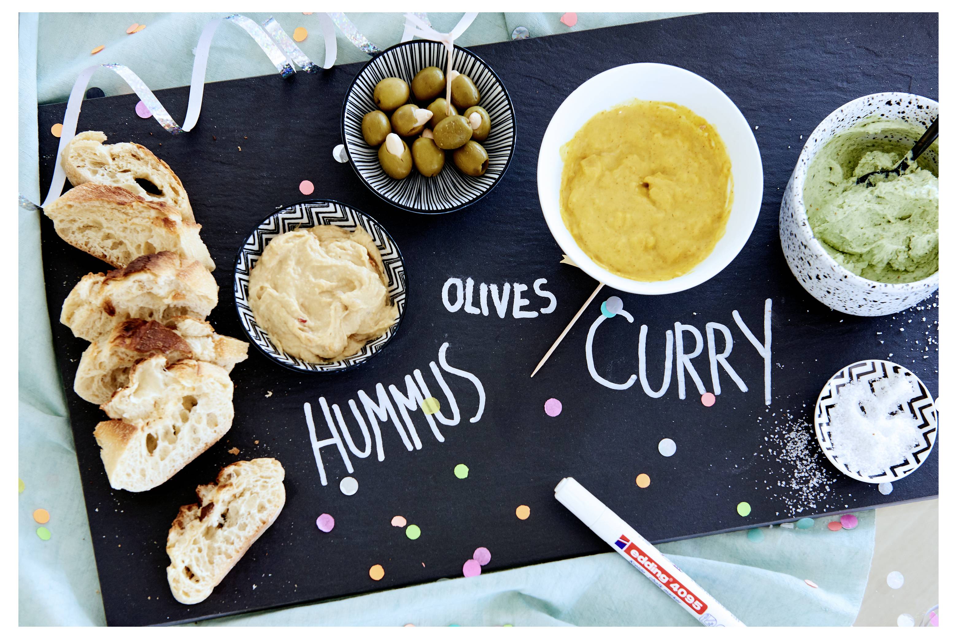 Draufsicht auf ein Snack-Board mit Brotscheiben, Oliven, Hummus, Curry-Dip und grünem Dip mit den Beschriftungen „HUMMUS", „OLIVEN" und „CURRY".