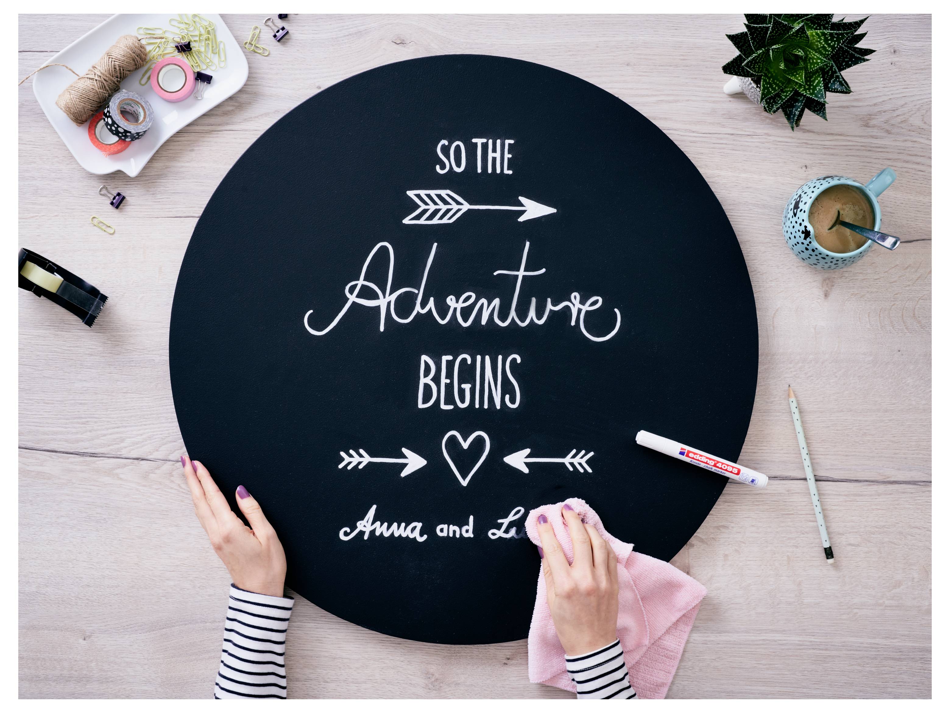 Hände arrangieren ein schwarzes rundes Tafelschild mit der Aufschrift "So the Adventure Begins" auf einem Schreibtisch mit Bastelmaterialien und einer Tasse Kaffee.