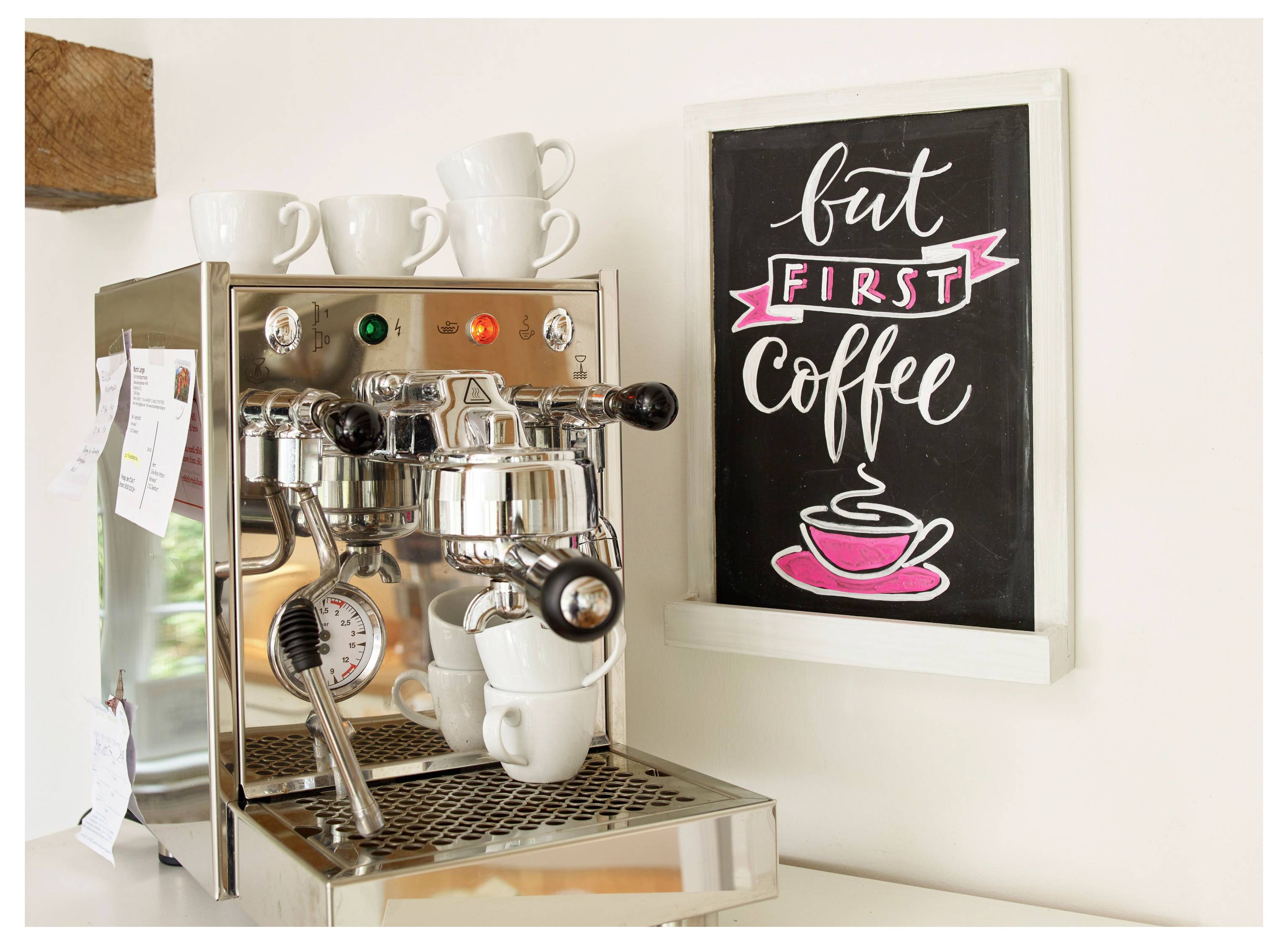 Espressomaschine mit Tassen neben einem gerahmten Schild mit der Aufschrift „aber ZUERST Kaffee" und einer Illustration einer rosa Tasse.