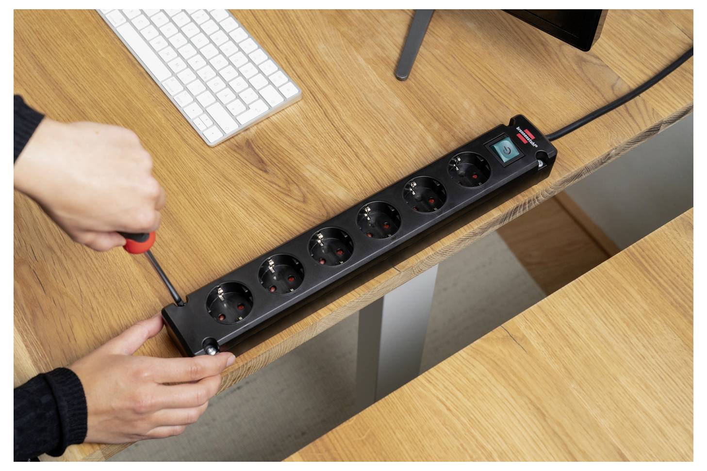 Hände installieren eine schwarze Steckdosenleiste mit mehreren Steckdosen unter einem Holzschreibtisch in der Nähe einer Tastatur und eines Monitors.