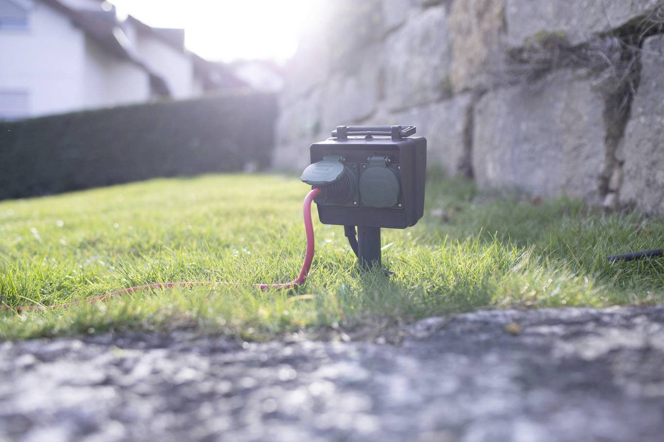 Eine Außensteckdose mit zwei Anschlüssen im Garten, teilweise von Gras verdeckt. Ein rotes Kabel ist angeschlossen. Licht scheint von hinten.