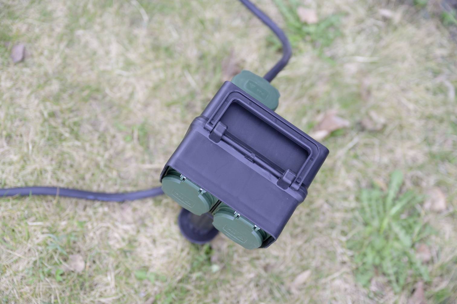 Eine Outdoor-Steckdose mit zwei grünen Buchsen und einem schwarzen Kabel liegt auf einer Rasenfläche.
