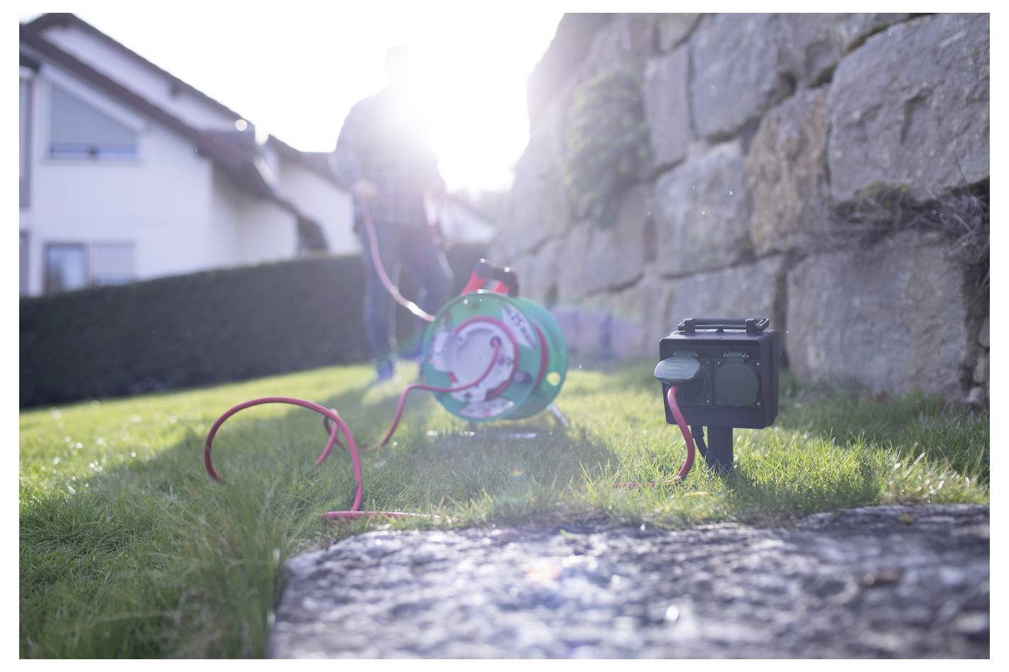 Person arbeitet im Freien mit einer grünen Schlauchrolle und einer schwarzen Pumpe oder einem Batteriepack auf dem Gras neben einer Steinmauer im hellen Sonnenlicht.