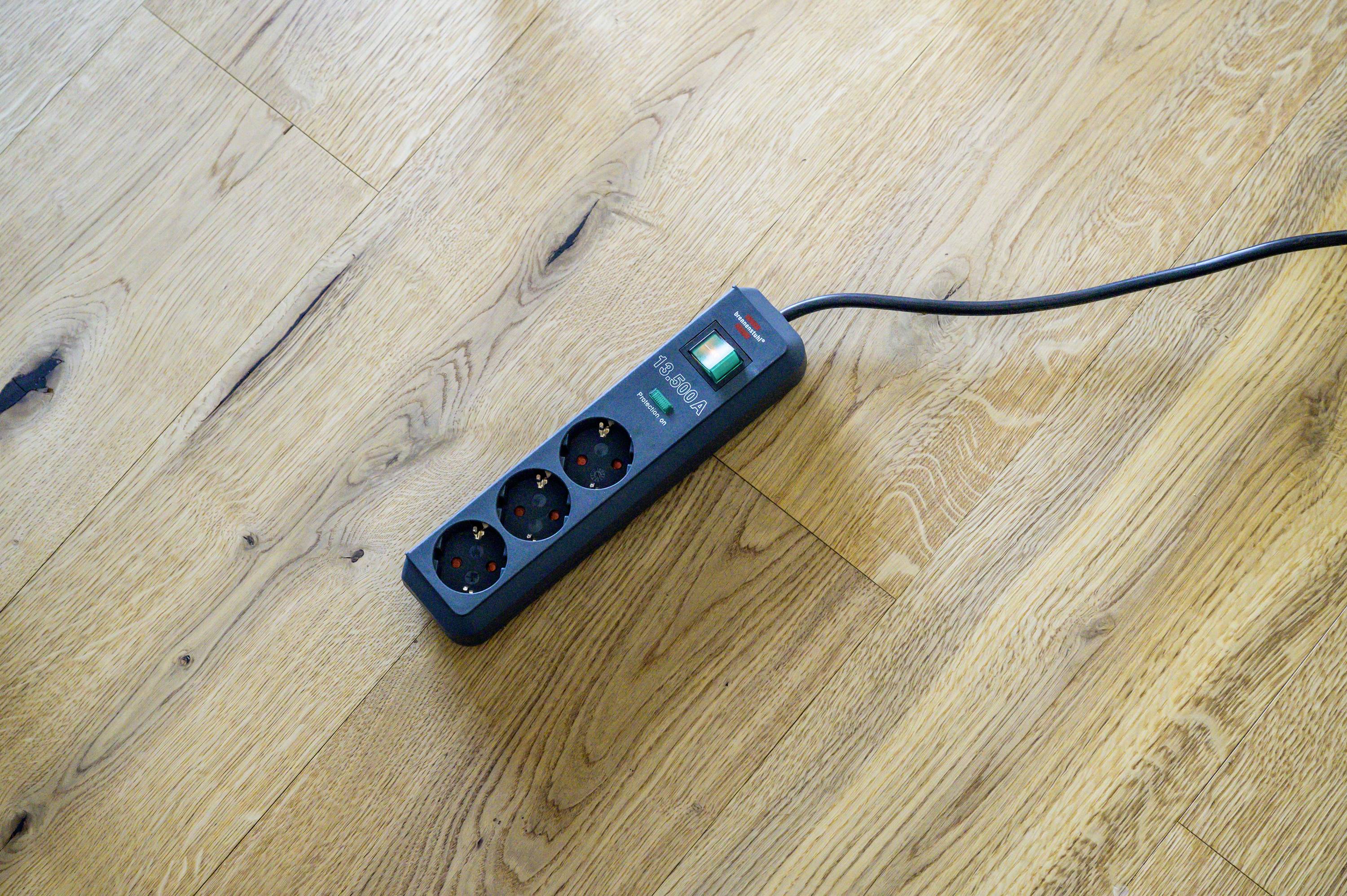 Drei Steckdosenleiste mit Schalter liegt auf einem Holzboden, verbunden mit einem Kabel.