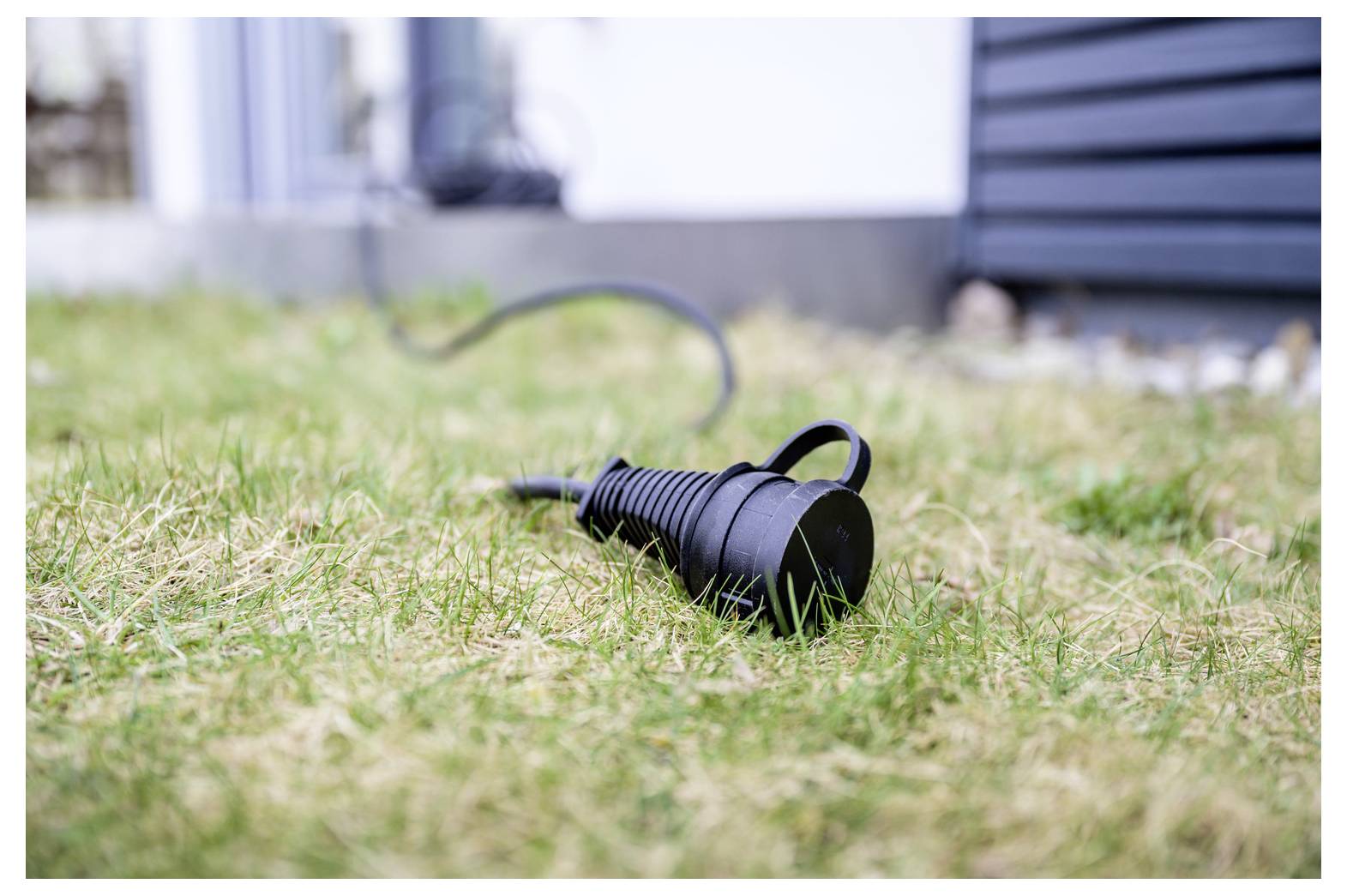 Schwarzes Außenkabel oder Schlauchverbindungsstück liegt auf Gras in der Nähe einer Gebäudeaußenseite, wobei sich das Kabel im Hintergrund erstreckt.