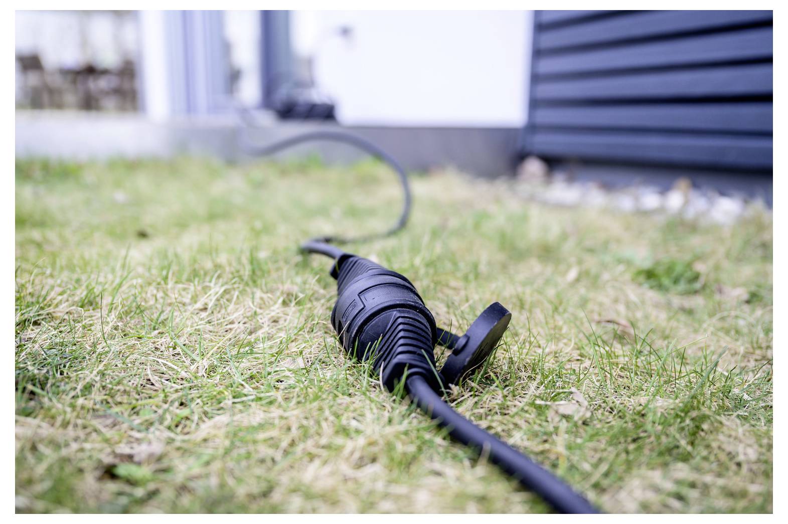 Nahaufnahme eines schwarzen elektrischen Steckerkabels, das auf dem Gras vor einem Haus liegt, wobei das Kabel zum Gebäude hin verläuft.