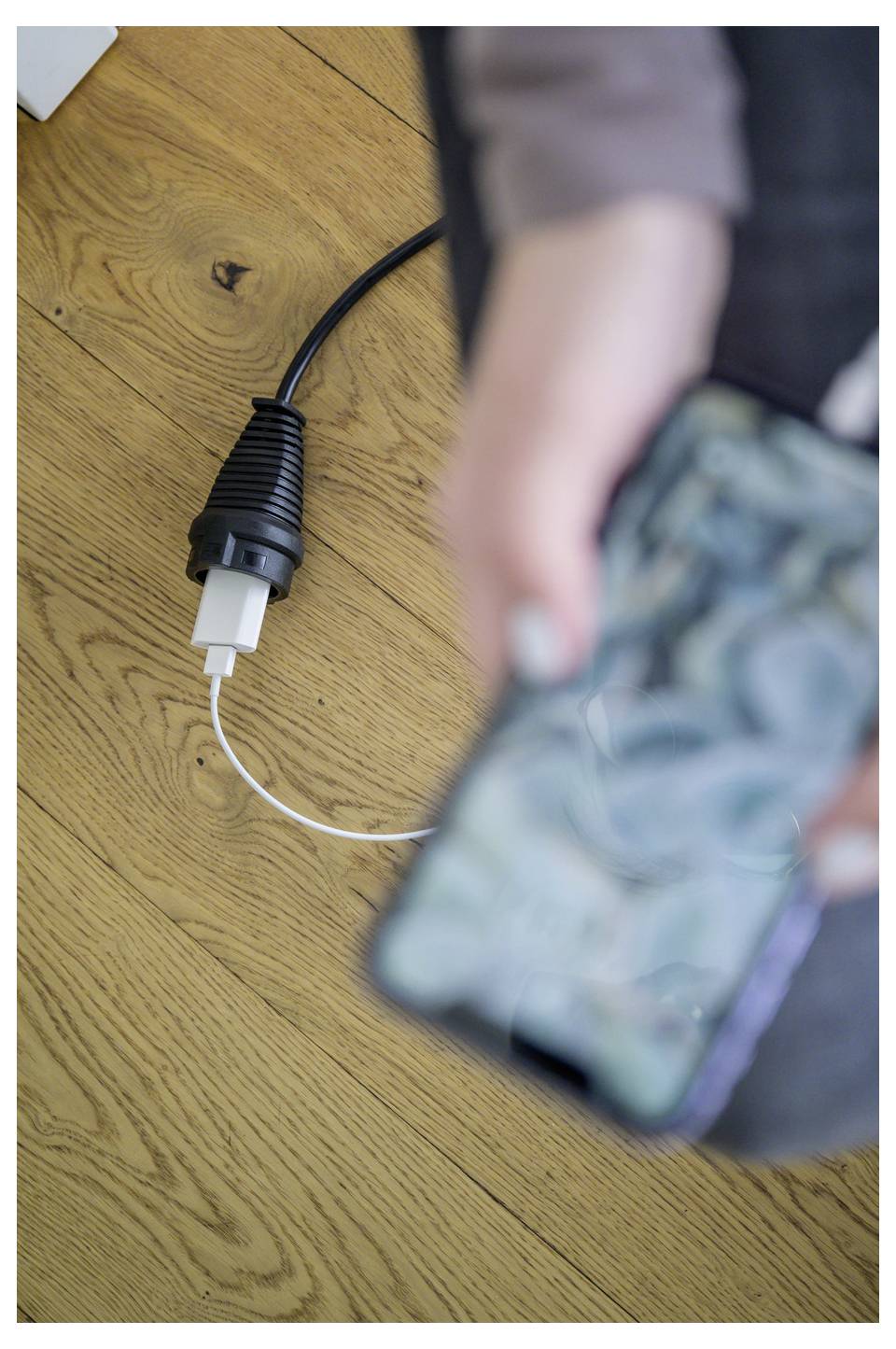 Nahaufnahme eines USB-Ladekabels auf einem Holzboden, während eine Person ein unscharfes Smartphone im Vordergrund hält.