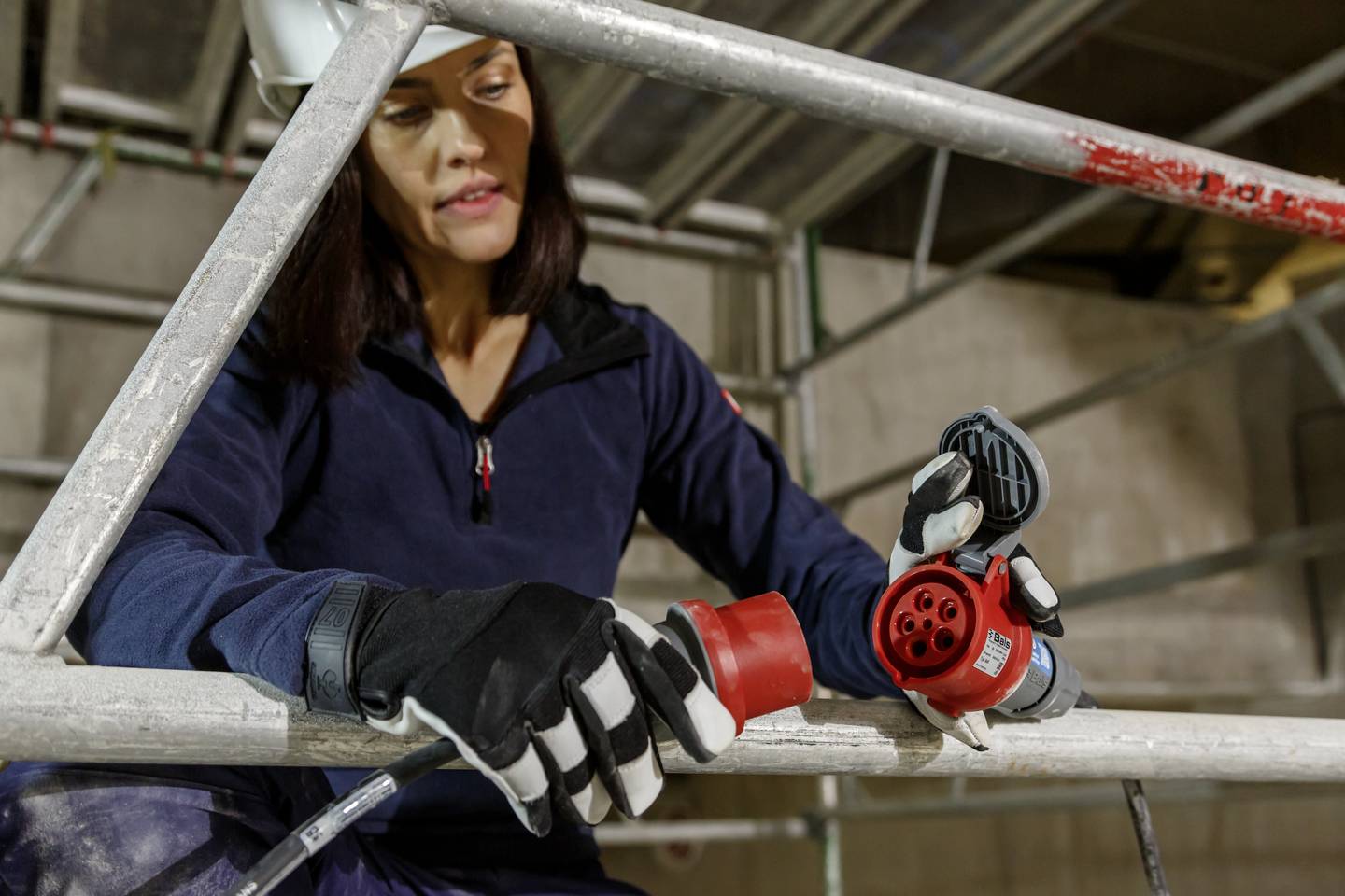 Eine Frau in Arbeitskleidung und Helm verbindet ein rotes Elektrokabel auf einer Baustelle. Sie trägt schwarze Handschuhe.