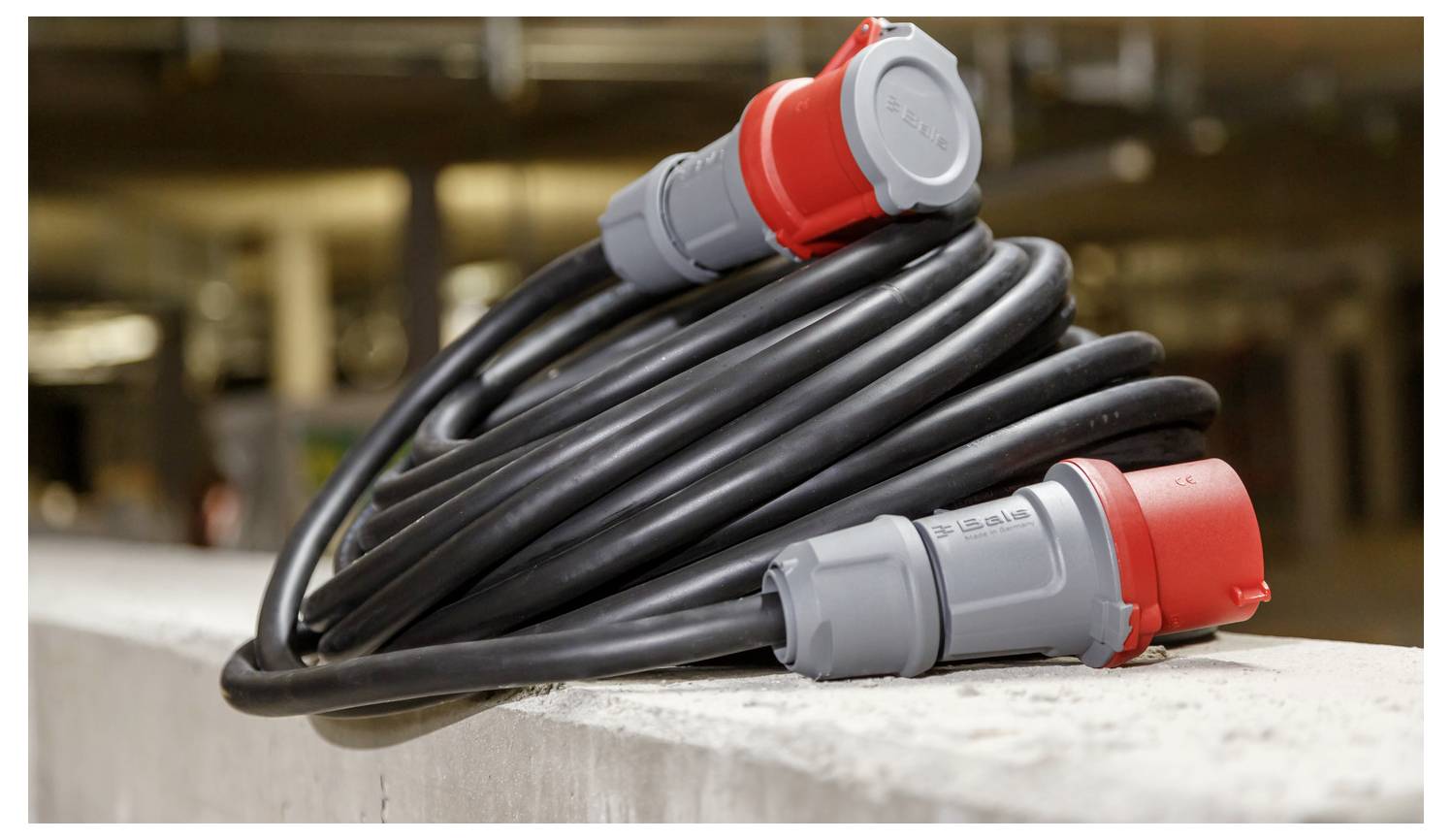 Aufgerolltes schweres Elektrokabel mit roten und grauen Industriesteckern auf einer Betonleiste ruhend.