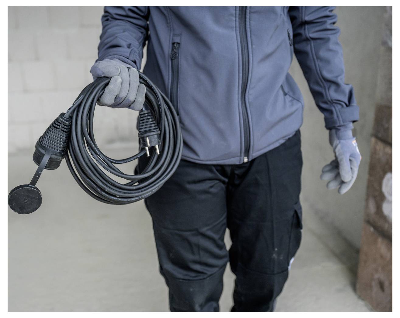 Person in Arbeitskleidung mit einer aufgerollten Stromleitung und Stecker in der Hand, mit einer runden schwarzen Werkzeugaufsatzvorrichtung, die daran hängt.