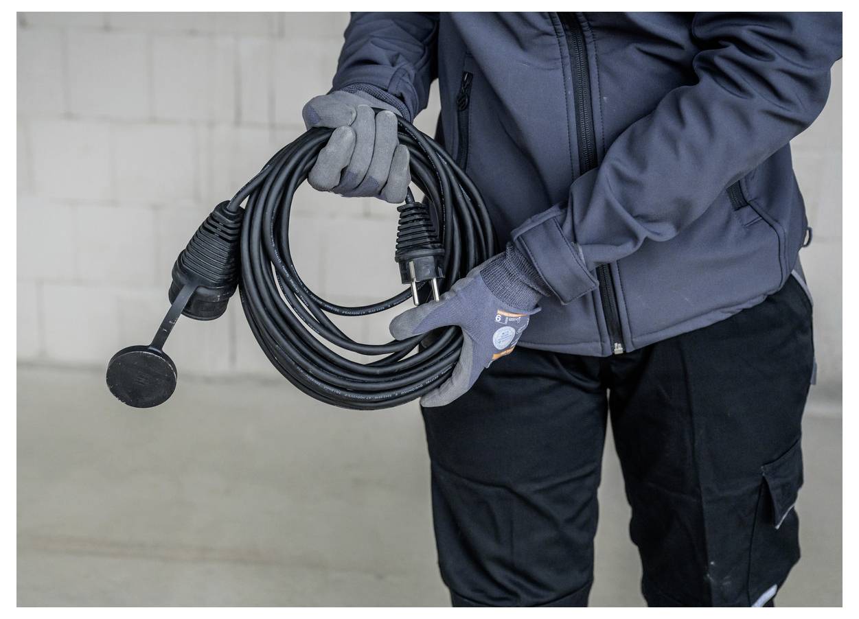 Handschuhgetragener Arbeiter hält ein aufgerolltes schwarzes Kabel mit einem Stecker und steckerartigen Mikrofon-ähnlichen Anschluss an einer schlichten Innenwand.