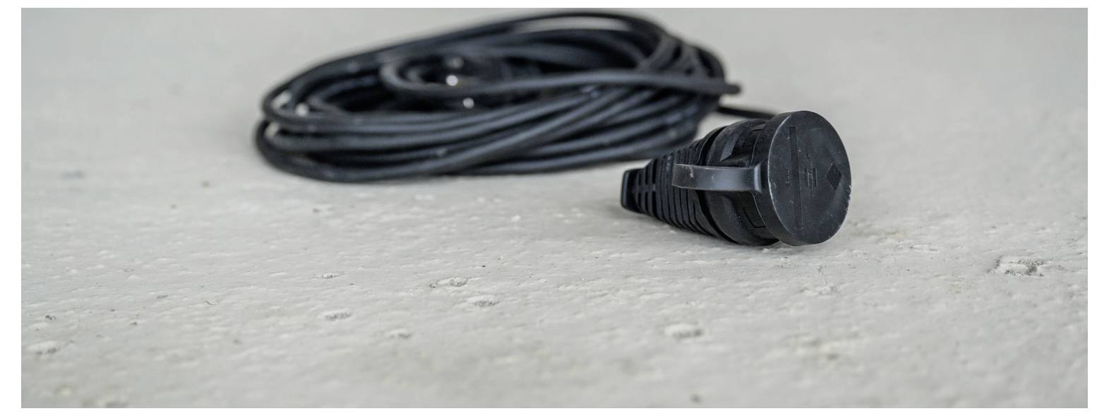 Nahaufnahme eines schwarzen Stromkabels oder einer schwarzen Elektroleitung mit einem Stecker, der aufgerollt auf einem hellgrauen Betonboden liegt.