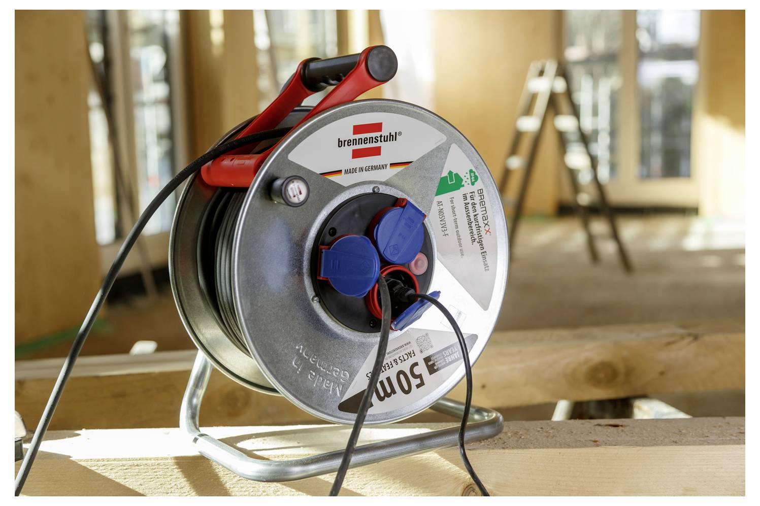 Eine Metallkabelrolle mit drei blauen Steckdosen und einem Stecker liegt auf dem Boden unter einer Holzkonstruktion und wird zur Verlängerung von Stromverbindungen verwendet.