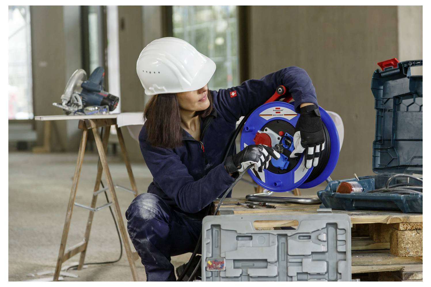 Elektrikerin in Schutzhelm knieend, um eine blaue Kabelrolle auf einer Werkbank in einer Werkstatt einzustellen, mit Werkzeugen in der Nähe.