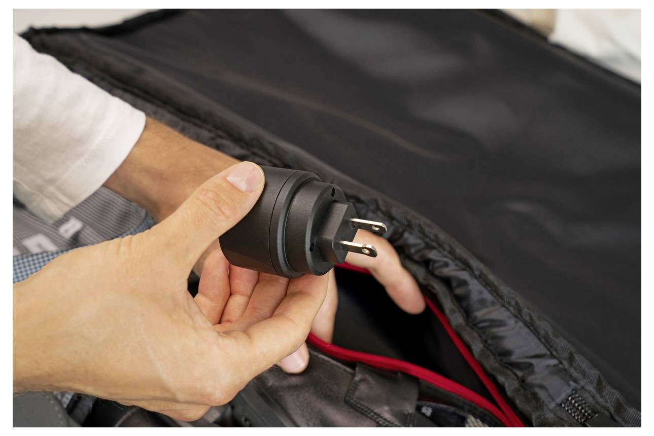 Hände halten einen schwarzen Stromstecker in der Nähe einer offenen schwarzen Tasche, wobei rote Fütterung sichtbar ist, was auf das Verpacken oder Anschließen eines Geräts hindeutet.