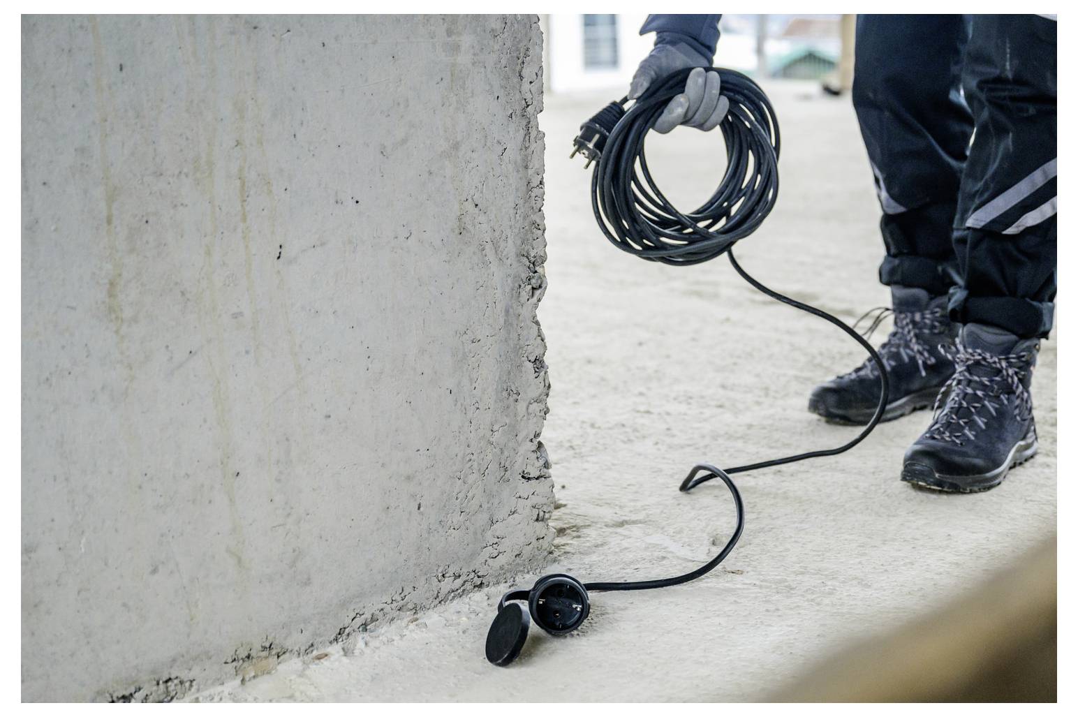 Arbeiter, der ein aufgerolltes Kabel neben einer Wand hält, wobei das Kabel zu einem Gerät auf dem Boden führt.