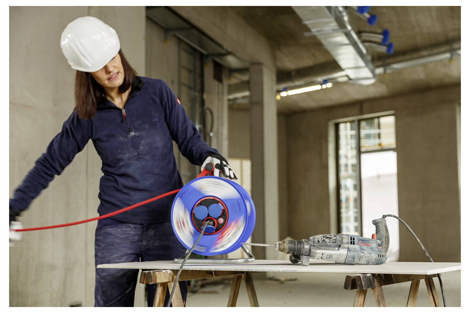 Bauarbeiter mit Schutzhelm, der eine blaue Kabelrolle neben einem Bohrer auf einer Werkbank im Innenraum eines Gebäudes handhabt.
