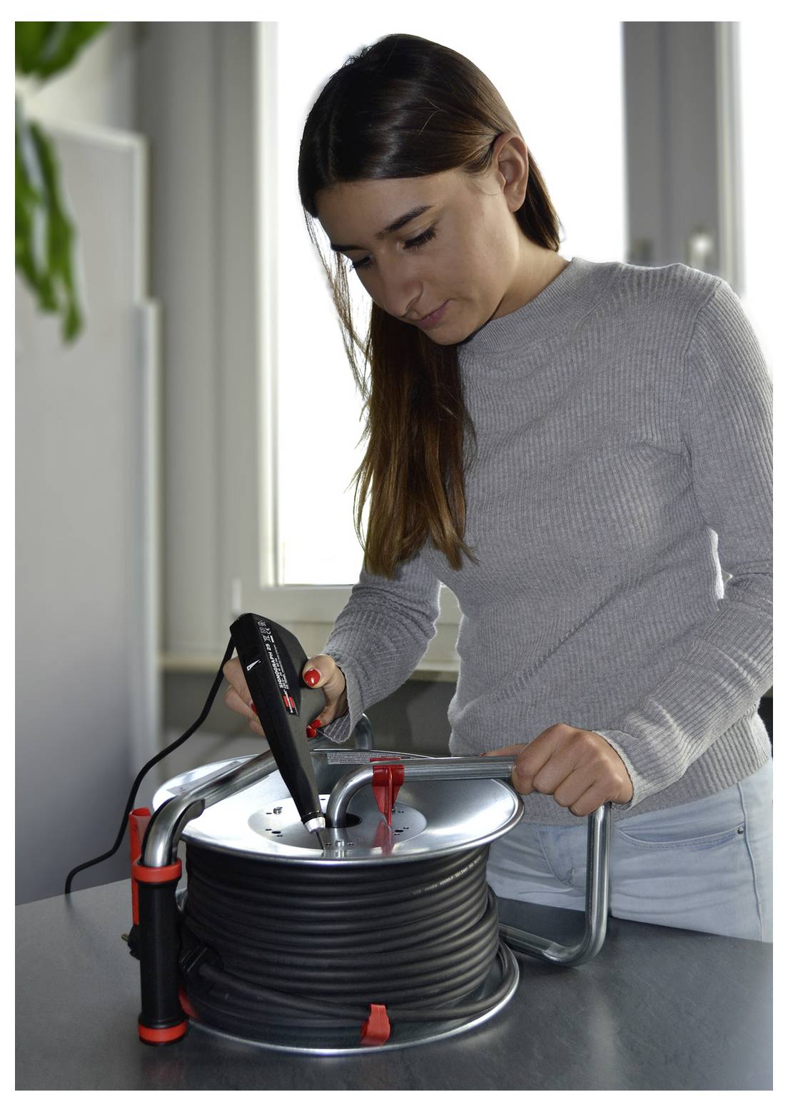 Frau, die einen Heißluftfön verwendet, um Schrumpffolie um eine Kabelspule auf einer Werkbank in Innenräumen zu schrumpfen.