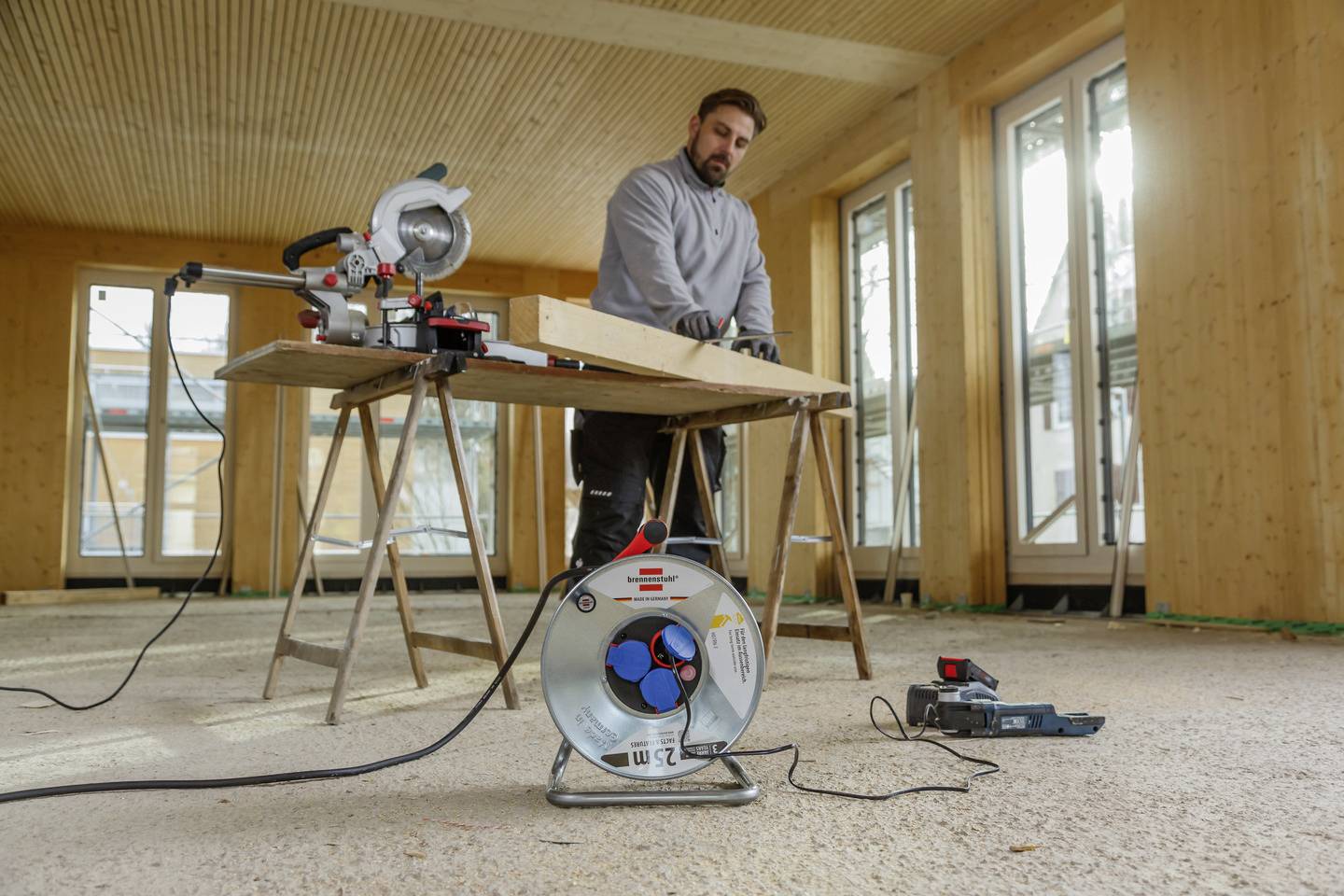 Eine Person arbeitet in einem Holzraum und verwendet eine Gehrungssäge auf einer Werkbank. Ein Verlängerungskabel und Elektrowerkzeuge liegen in der Nähe auf dem Boden.