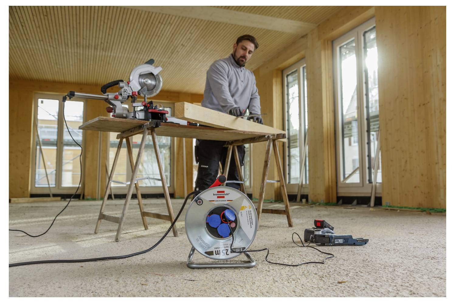 Ein Mann bedient eine Gehrungssäge in einem teilweise fertiggestellten Zimmer, während er ein langes Holzbrett zuschneidet; eine Kabelrolle und Werkzeuge liegen auf dem Boden.