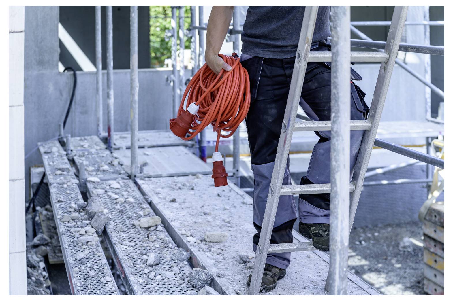 Ein Bauarbeiter hält ein oranges Verlängerungskabel in der Hand, während er auf einem Gerüst neben einer Leiter auf einer Baustelle steht.