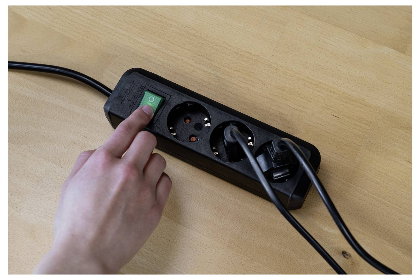 Eine Hand schaltet eine schwarze Steckdosenleiste auf einem Holzschreibtisch ein, mit eingesteckten Kabeln, die mit den Steckdosen verbunden sind.