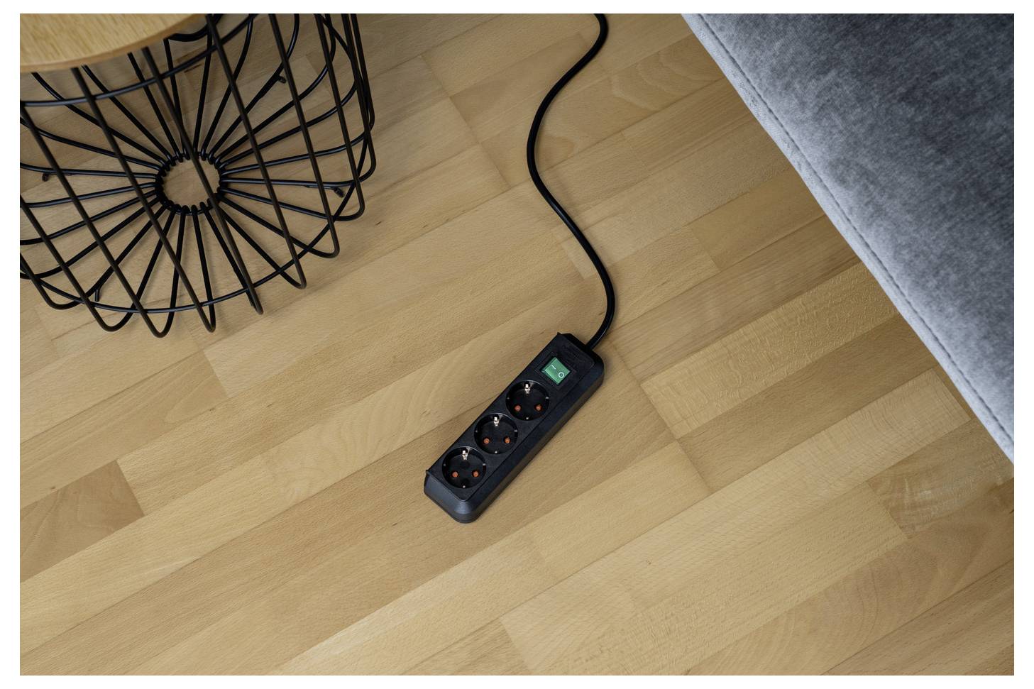 Steckerleiste mit drei Steckdosen und einem grünen Netzschalter auf einem Holzboden neben einem Sofa und einem Drahtkorb