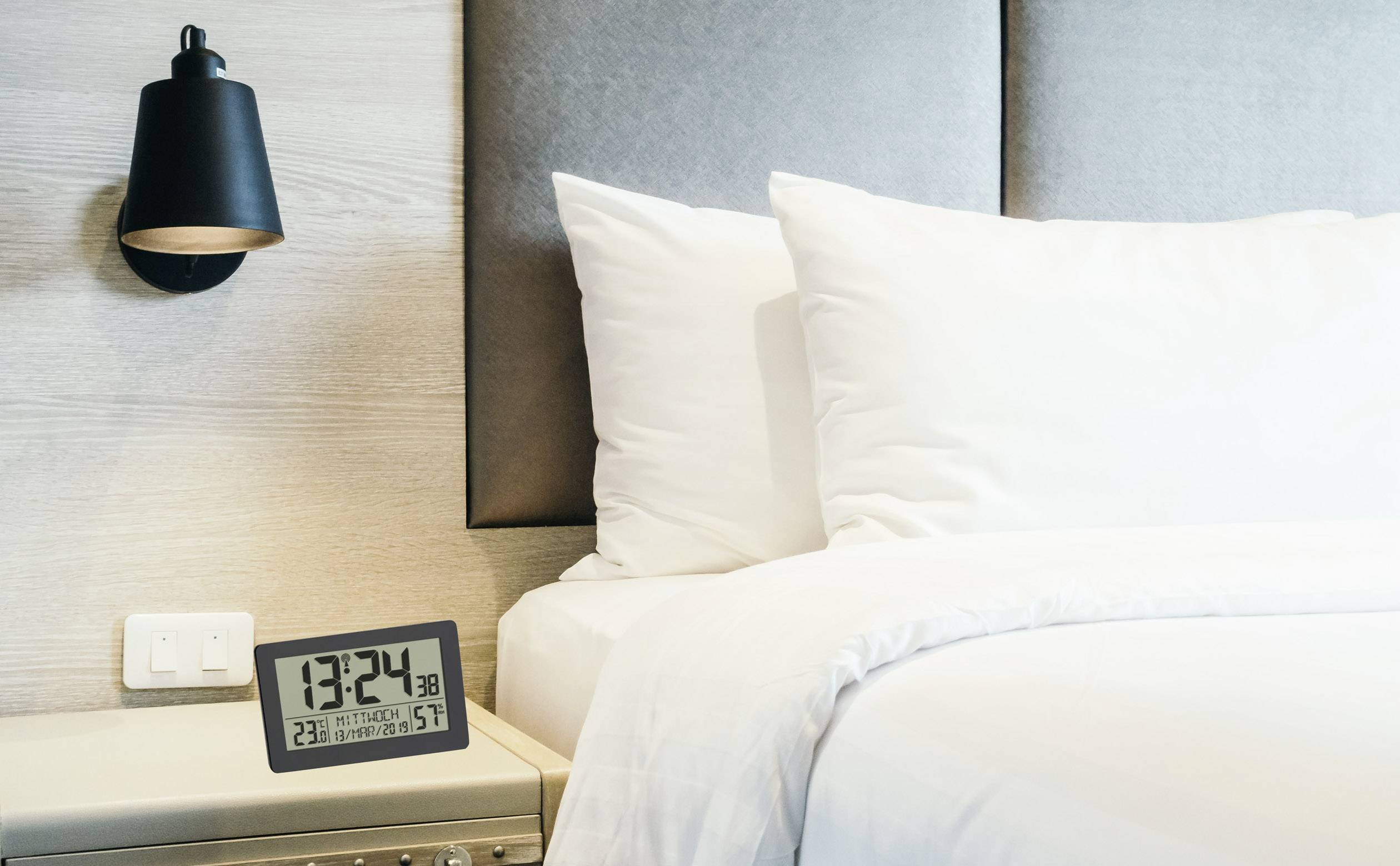 Digitaluhr zeigt 13:24 auf Nachttisch neben Bett mit weißen Kissen und einer Decke; moderne Lampe an der Wand.