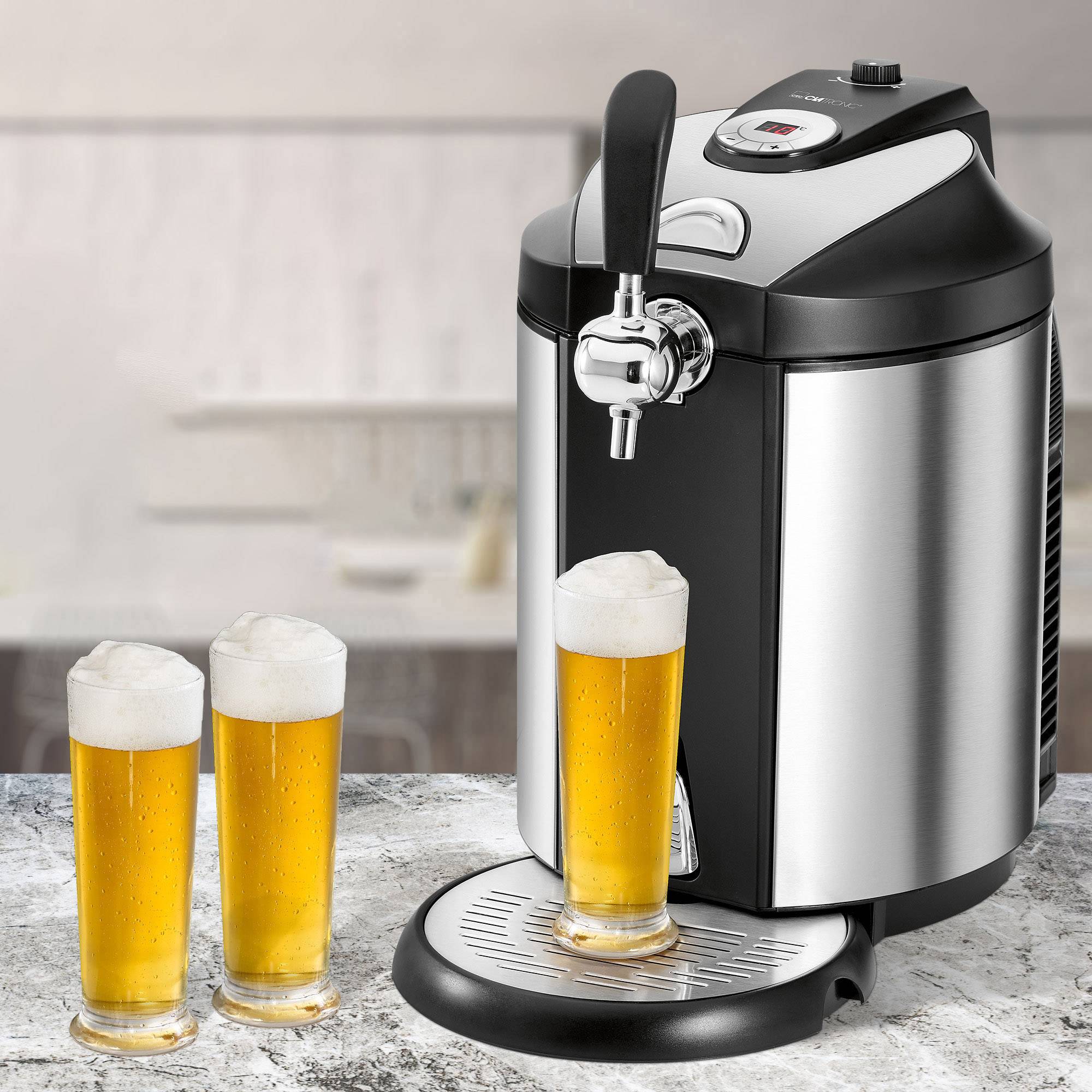 Eine silberne Bierzapfanlage steht auf einem Tisch. Drei Gläser gefüllt mit hellem Bier und Schaum stehen daneben. Im Hintergrund ist eine unscharfe Küche.