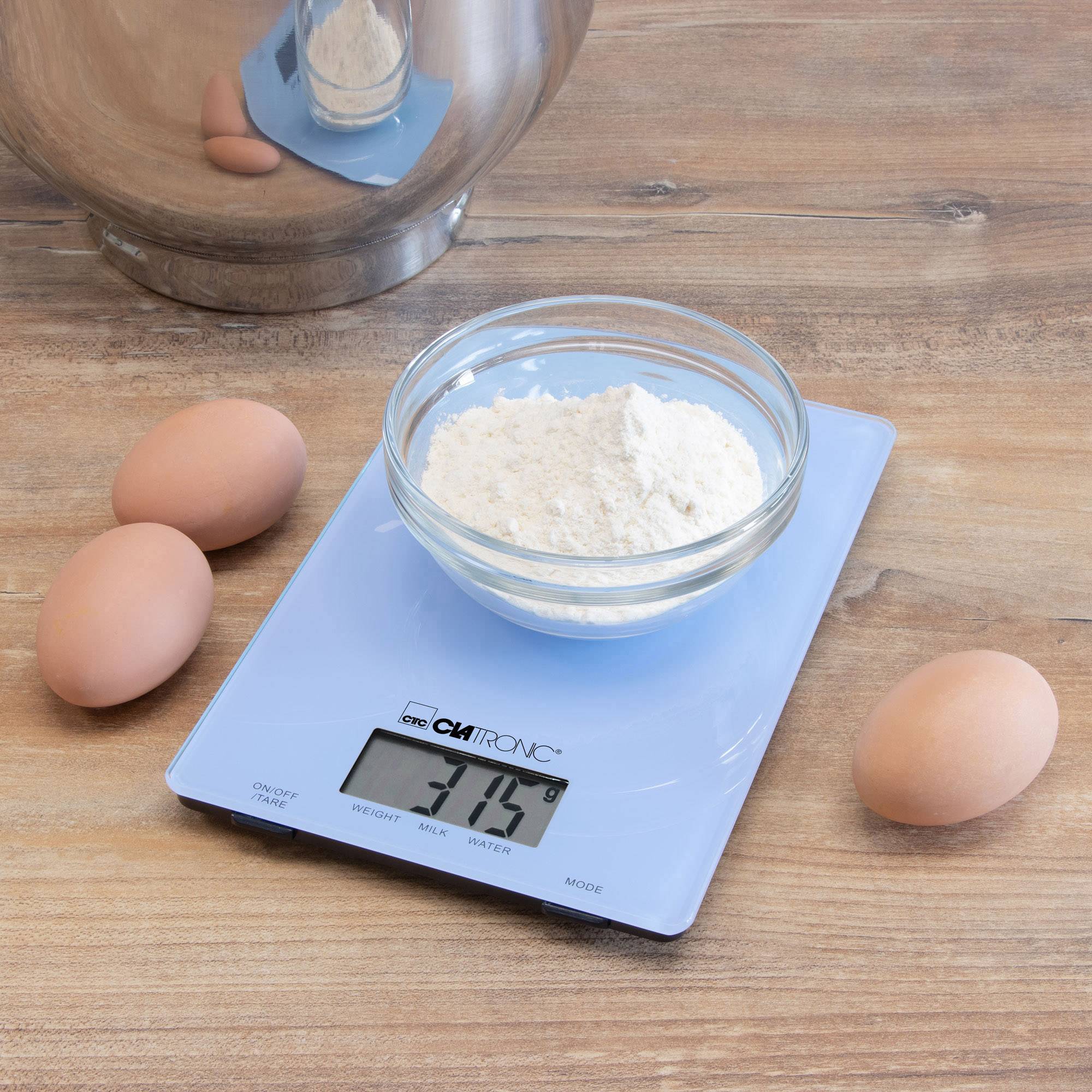 Eine digitale Küchenwaage zeigt 315 g Mehl in einer Glasschale an. Daneben liegen vier braune Eier auf einer Holzoberfläche.