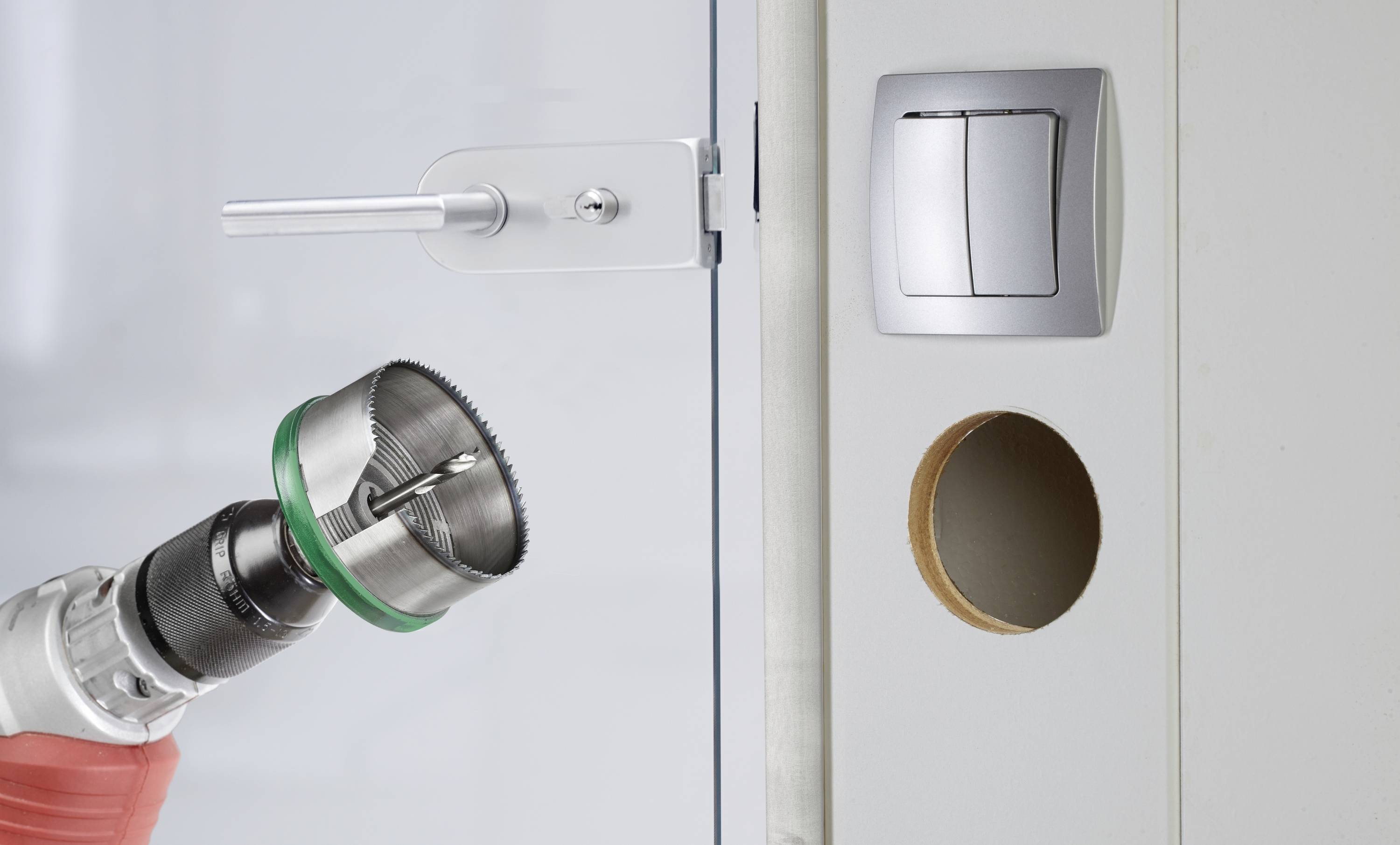 Bohrmaschine mit Lochsäge, die ein rundes Loch in eine Tür bohrt. Daneben ein Lichtschalter.