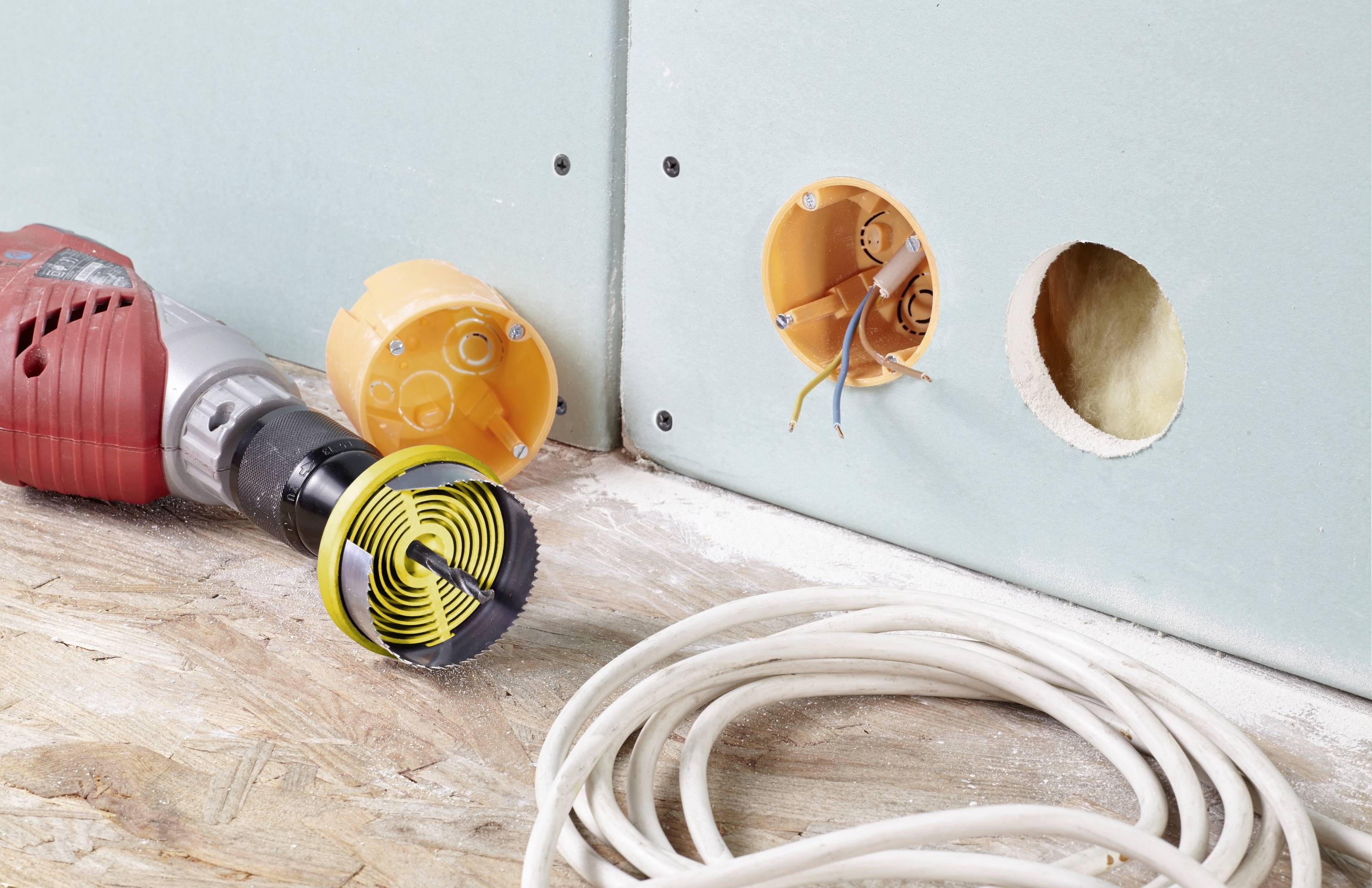 Bohrmaschine mit Lochsäge liegt vor einer Trockenbauwand. Zwei Wanddosen sind sichtbar. Kabel sind bereit zur Elektroinstallation.