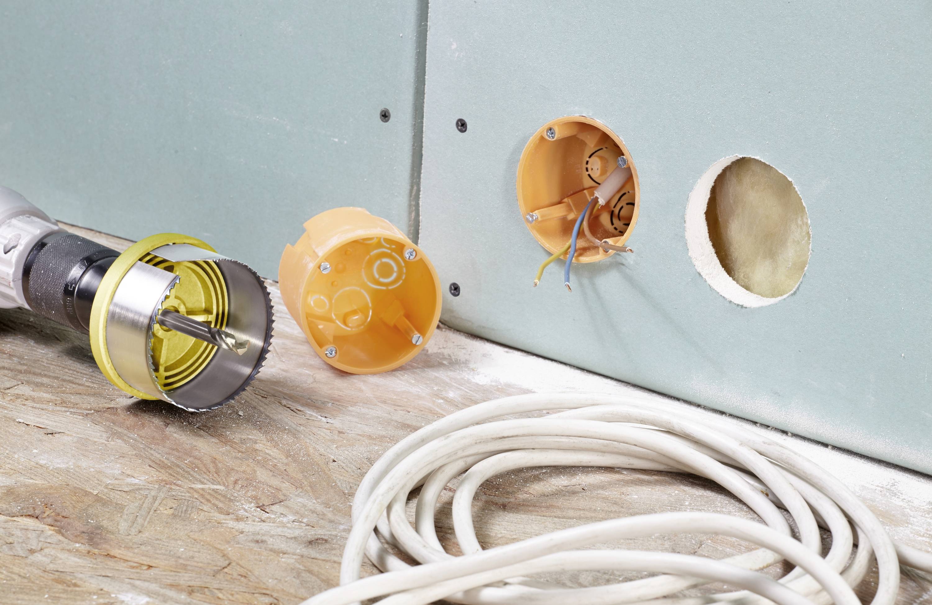 Bohrmaschine mit Lochsäge und Elektroinstallationsdosen an einer Trockenbauwand; Stromkabel auf dem Boden. Baustellenszene.