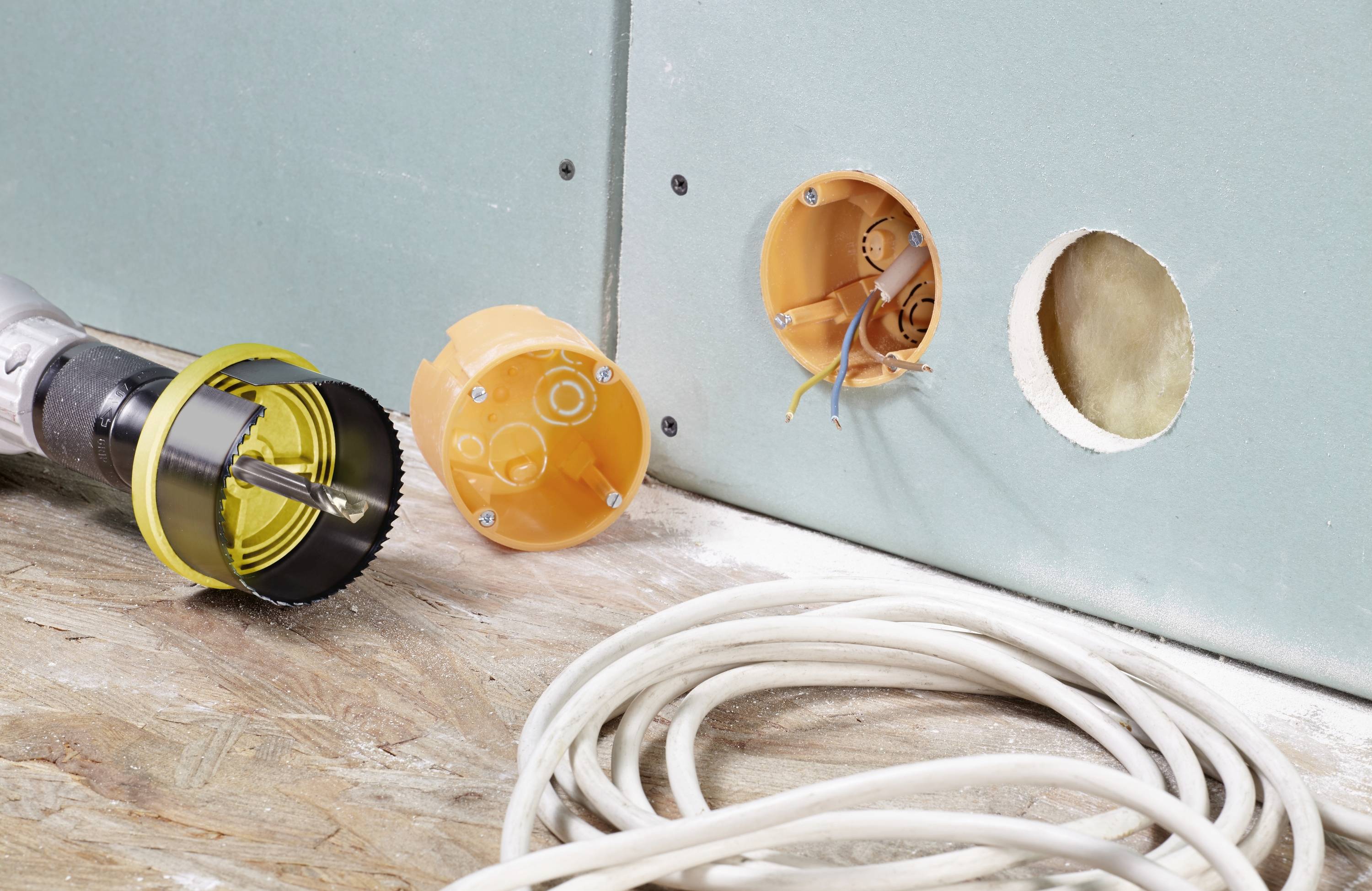 Ein Werkzeug mit Bohrkrone ist neben einer Trockenbauwand mit gebohrtem Loch zu sehen. Daneben liegen Kabel und eine orangefarbene Verteilerdose.