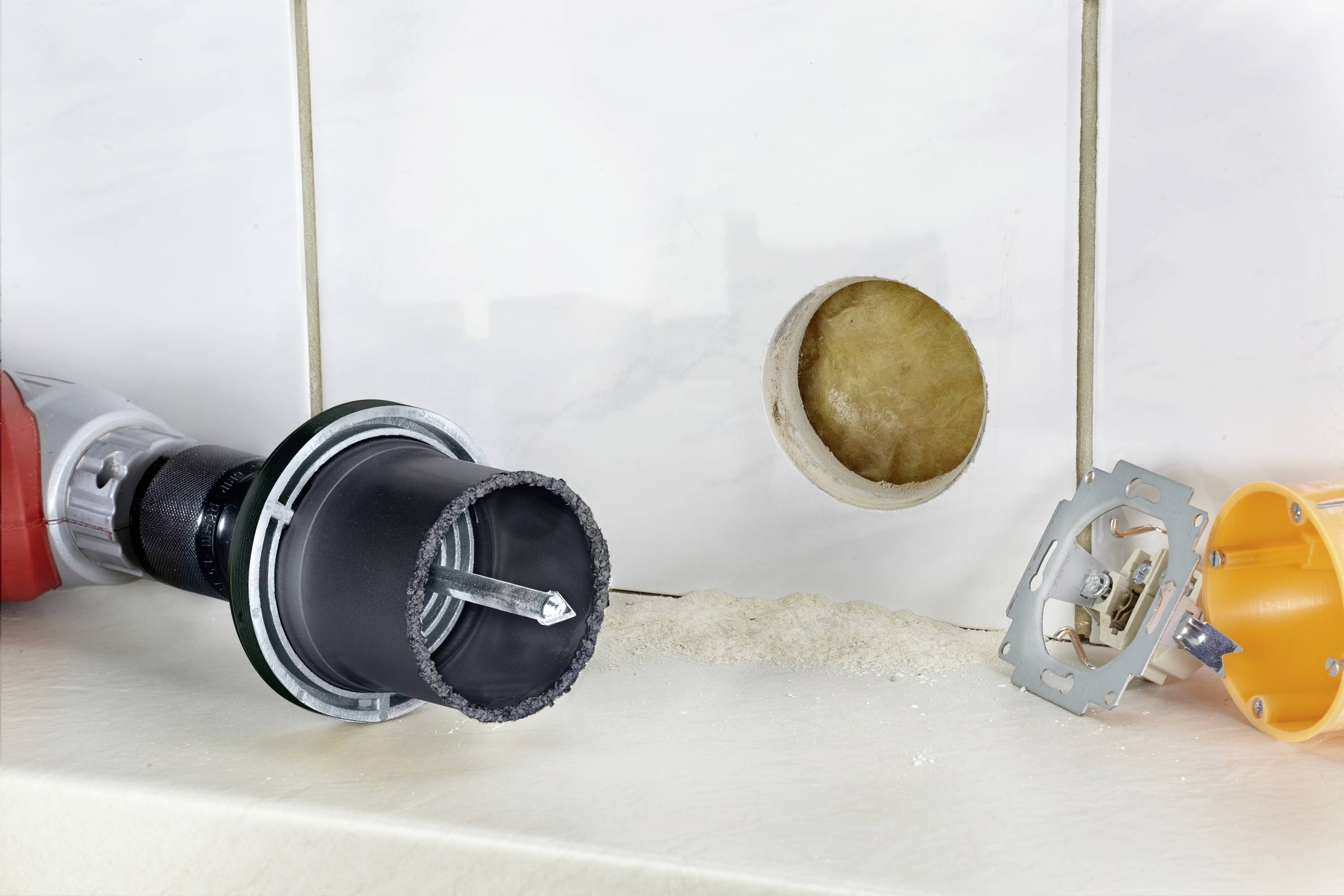 'Werkzeug zum Fliesenbohren neben einer gebohrten Fliese und Steckdosenteilen'; zeigt Heimwerkerbedarf und Renovierungsprozess.