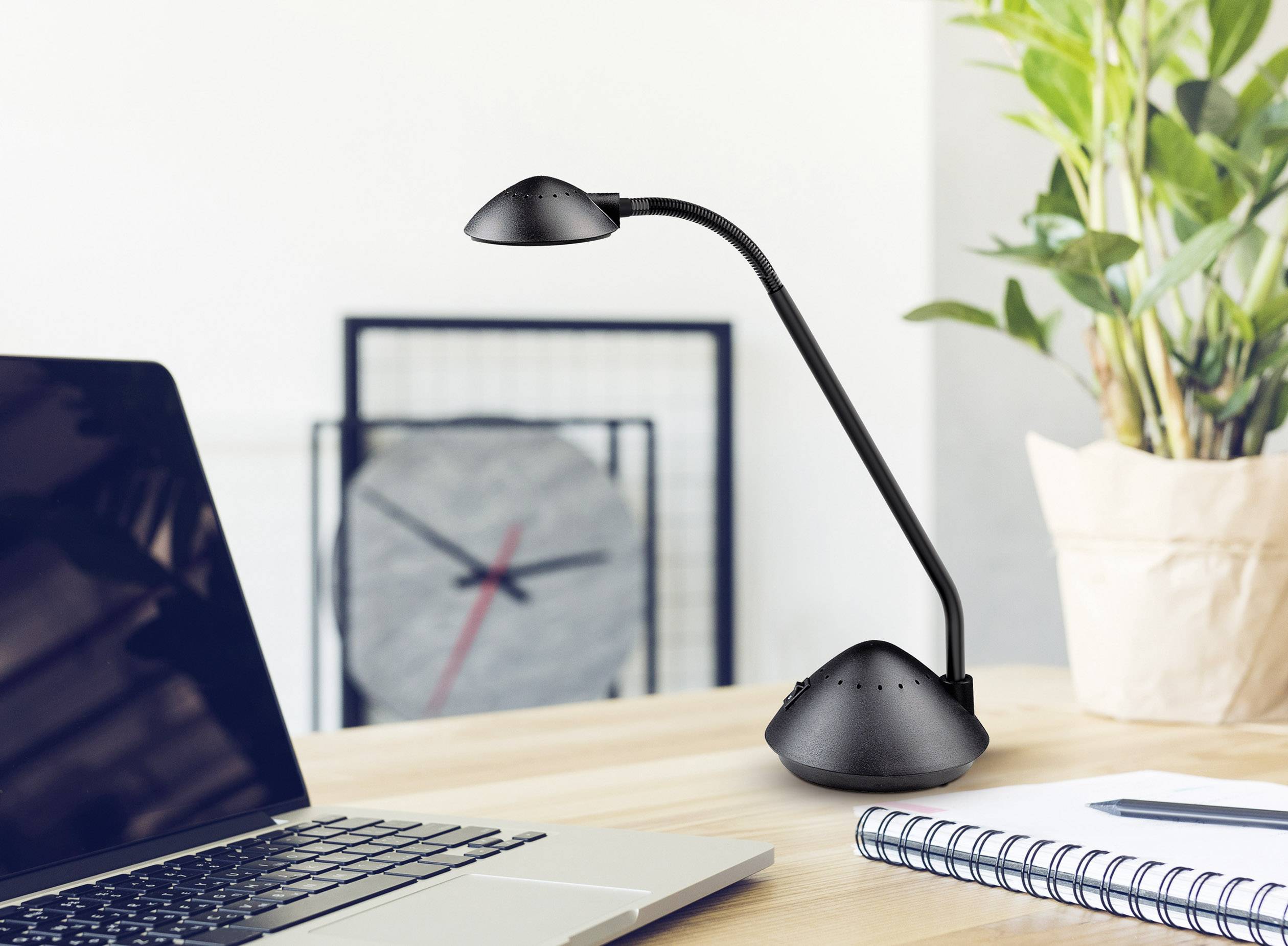 Ein moderner Schreibtisch mit einem Laptop, einer schwarzen Tischlampe und einer Topfpflanze. Im Hintergrund ist eine Wanduhr sichtbar.