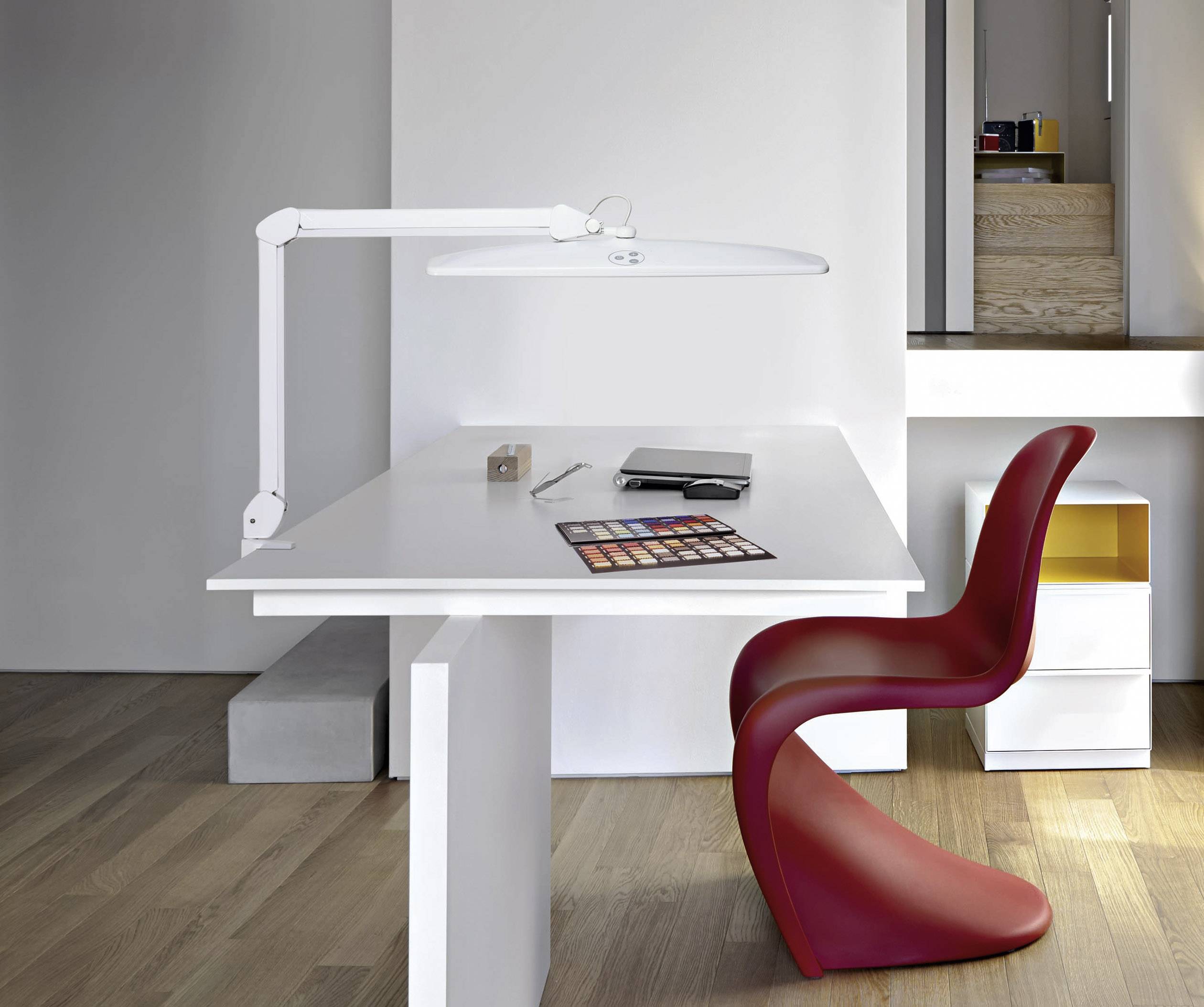 Ein moderner Arbeitsbereich mit weißem Tisch, rotem Stuhl und Schreibutensilien auf Holzboden; eine stilvolle Lampe beleuchtet den Tisch.