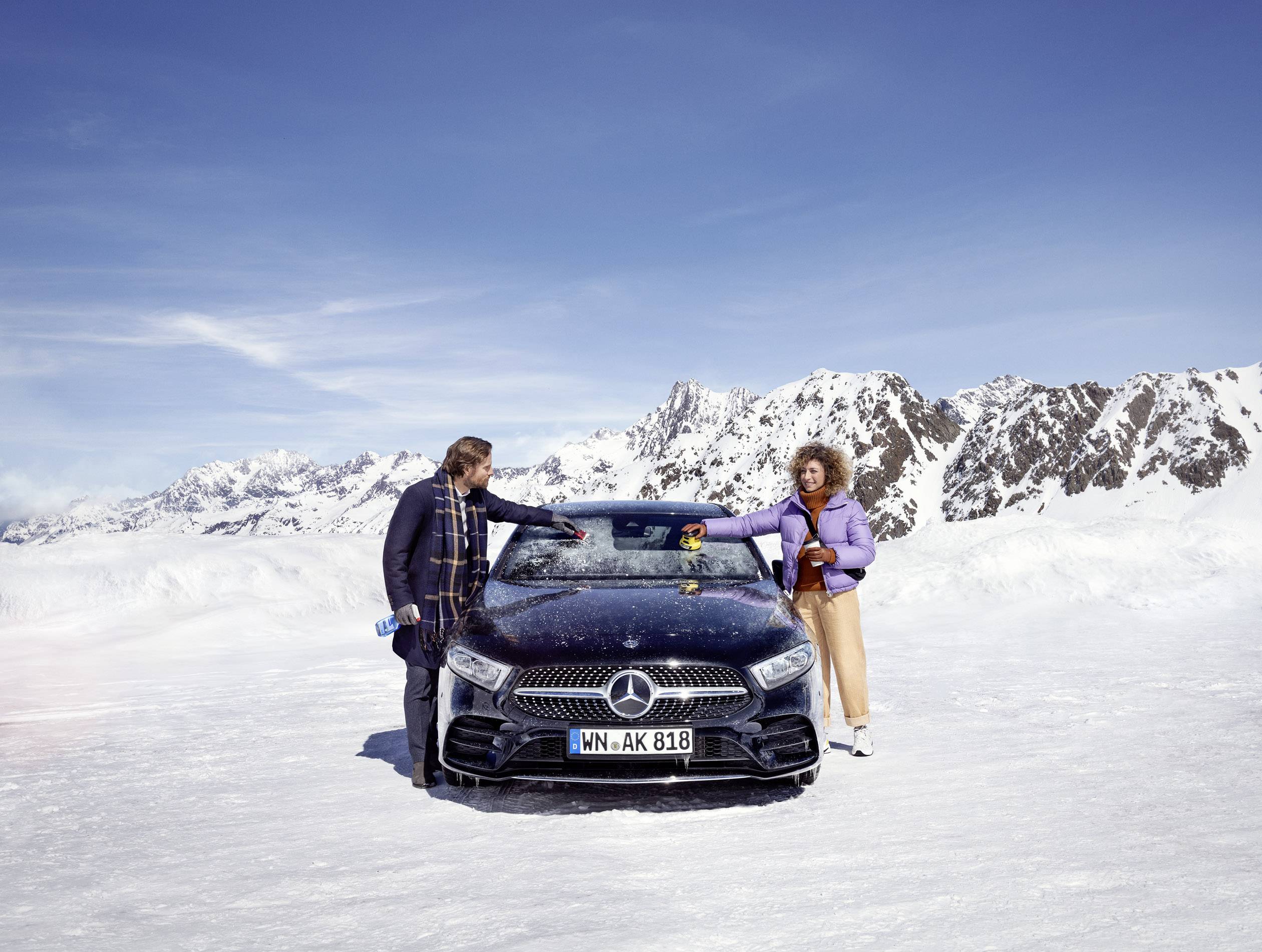 Zwei Personen stehen vor einem Mercedes auf einer verschneiten Berglandschaft. Eine Person trägt eine lila Jacke, die andere gestreifte Kleidung.
