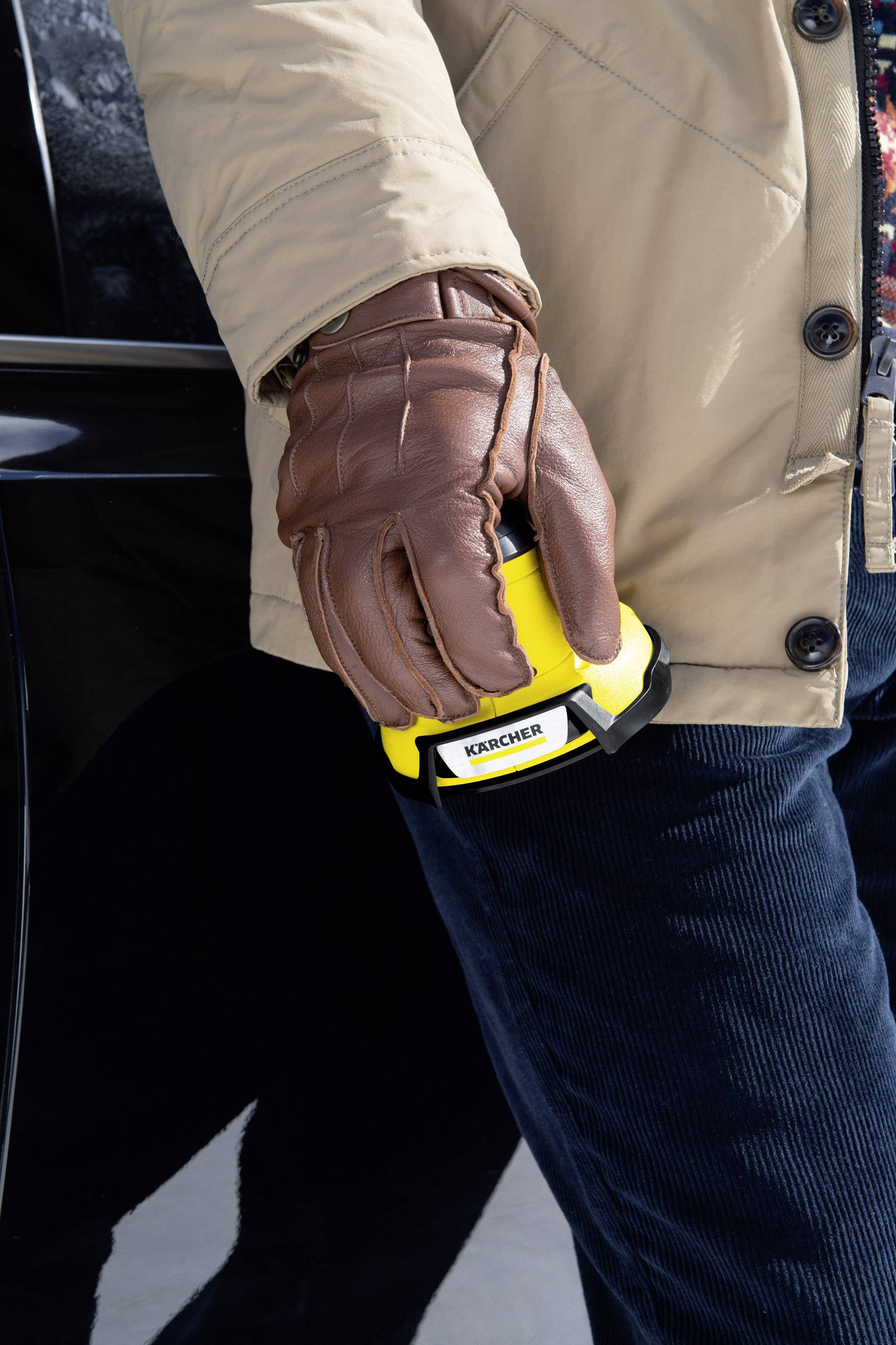 Eine Person trägt braune Handschuhe und hält ein gelbes Gerät mit dem Aufdruck 'Kärcher' neben einem schwarzen Auto.