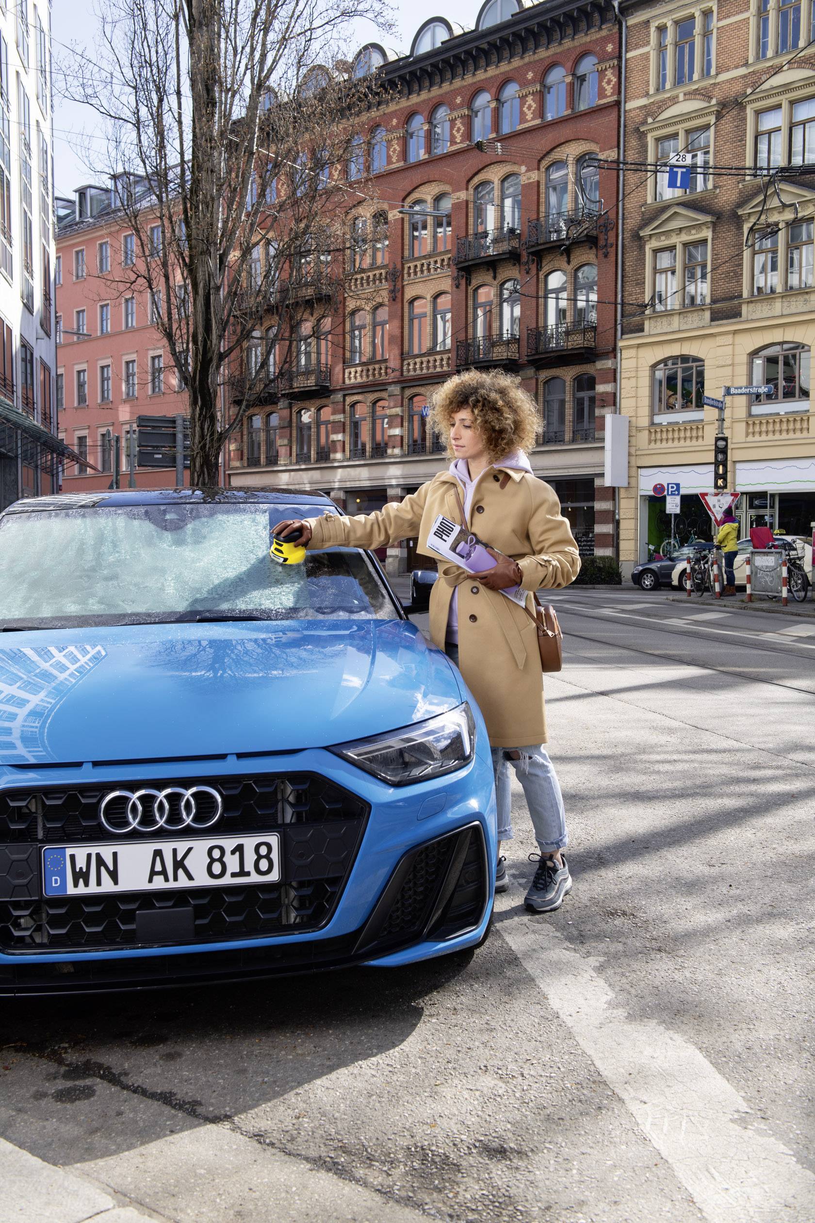 Eine Person entfernt Eis von der Windschutzscheibe eines blauen Autos mit einem Eiskratzer. Im Hintergrund sind Gebäude einer Stadt zu sehen.