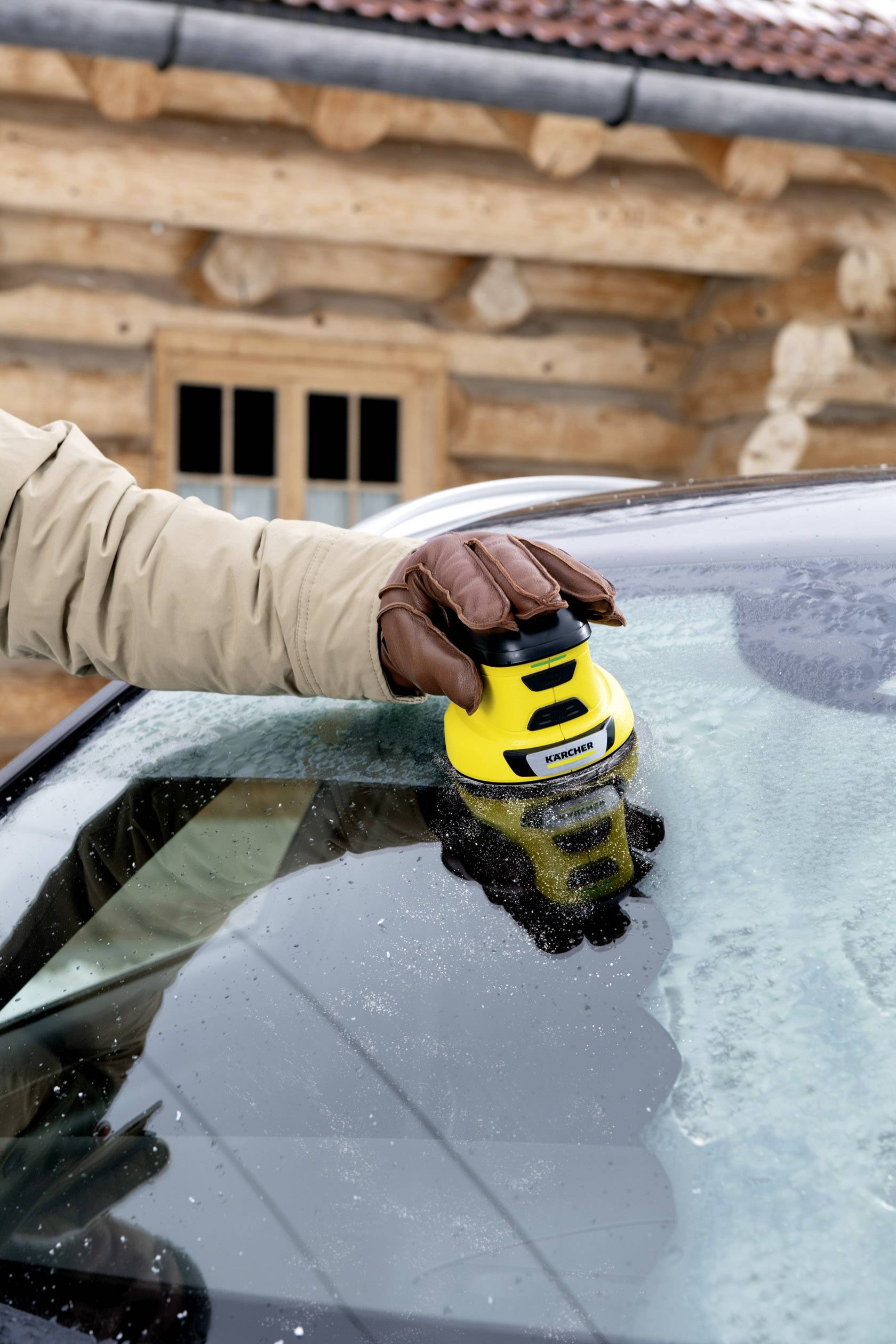 Person entfernt Eis von einer Autoscheibe mit einem gelben Eiskratzer vor einem Holzhaus im Winter.