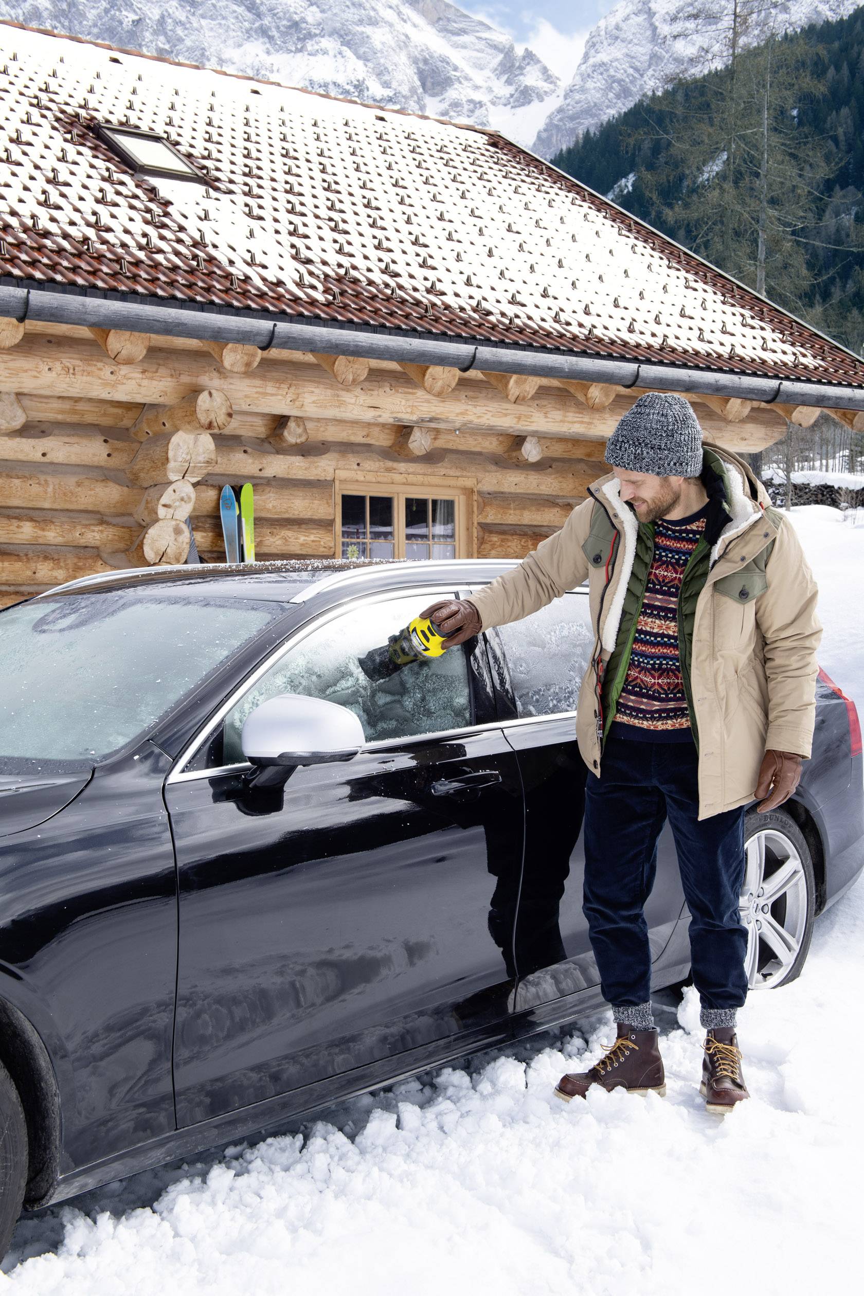 Ein Mann steht im Schnee neben einem Auto und entfernt Eis von der Windschutzscheibe mit einem Eiskratzer, ein Holzhaus im Hintergrund.